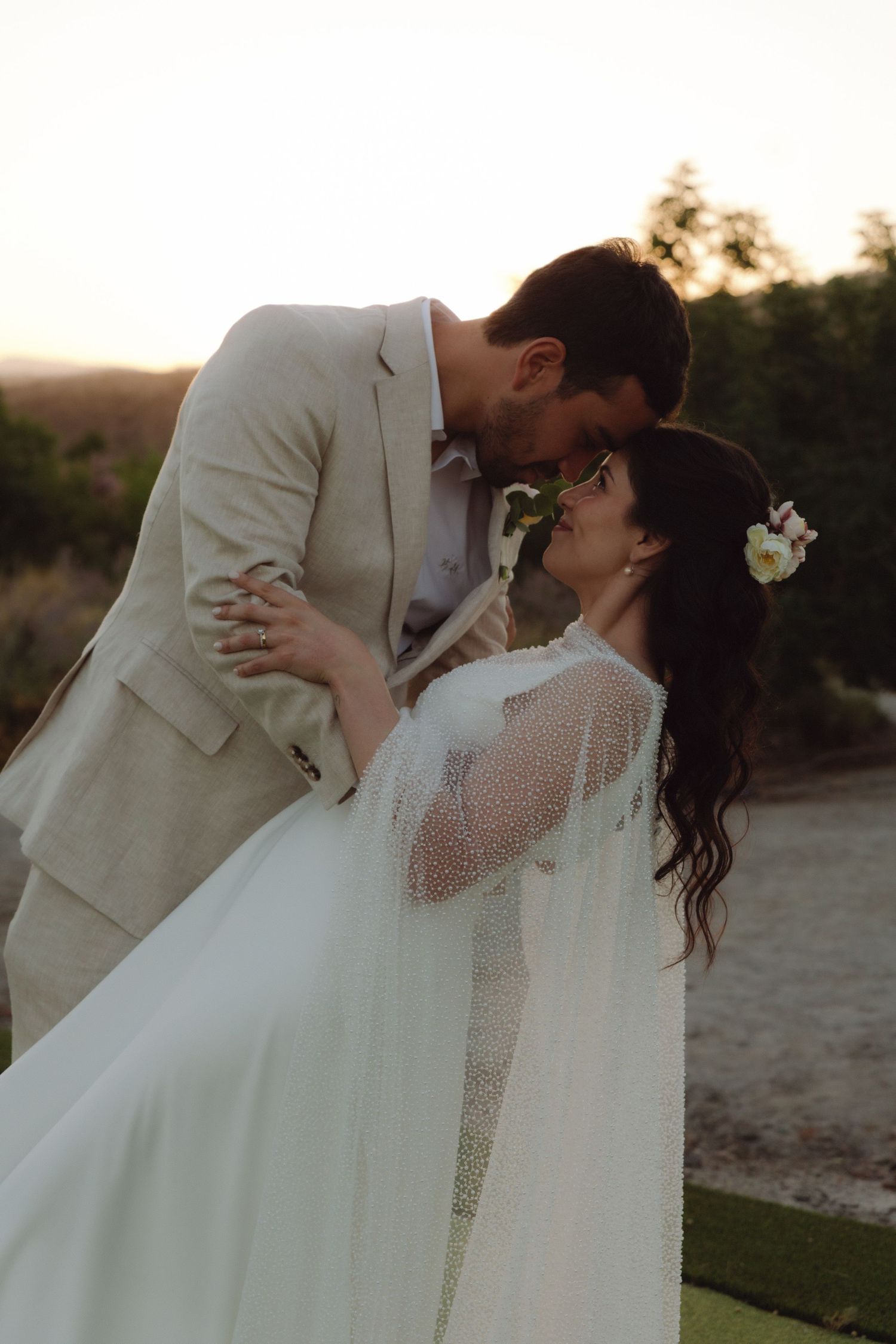 Groom leaning in to kiss bride wearing floral hair accessory at outdoor sunset wedding.
