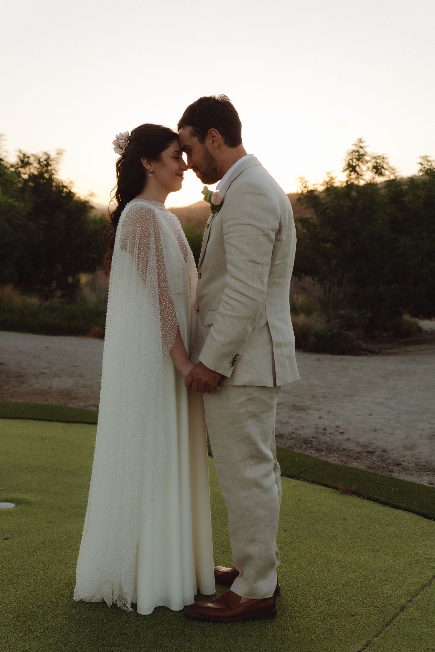 Bride and groom touching foreheads tenderly while holding hands at golden hour wedding.