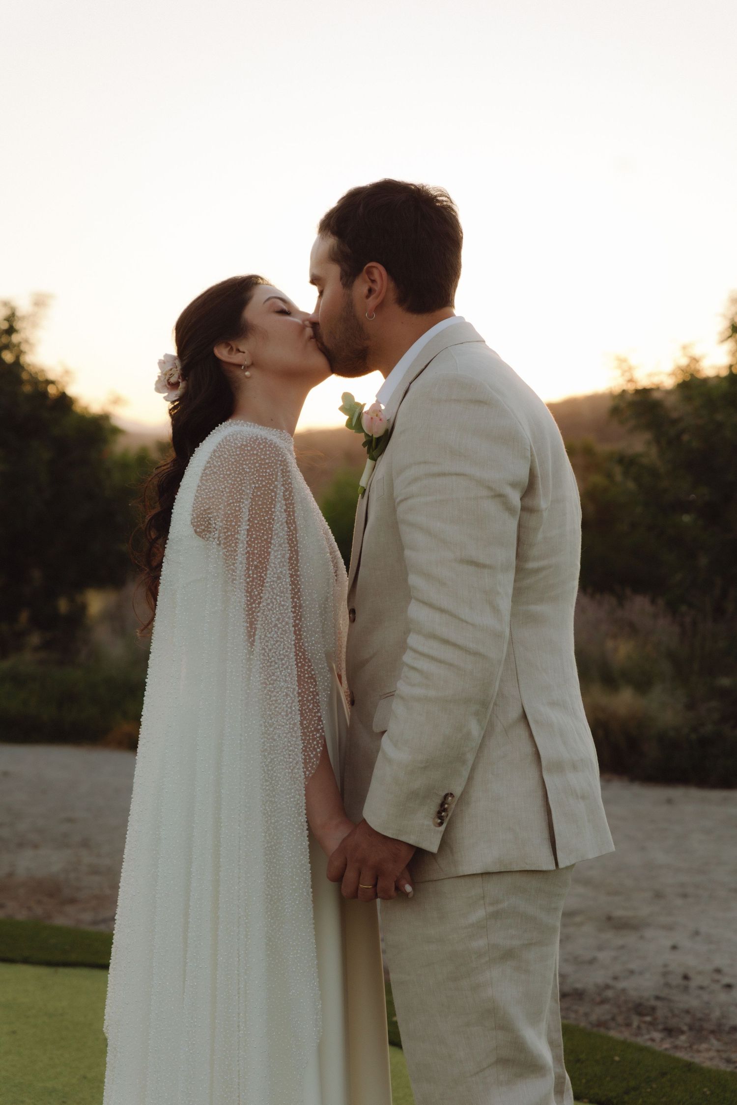 Bride and groom sharing a romantic kiss while holding hands at outdoor sunset wedding.