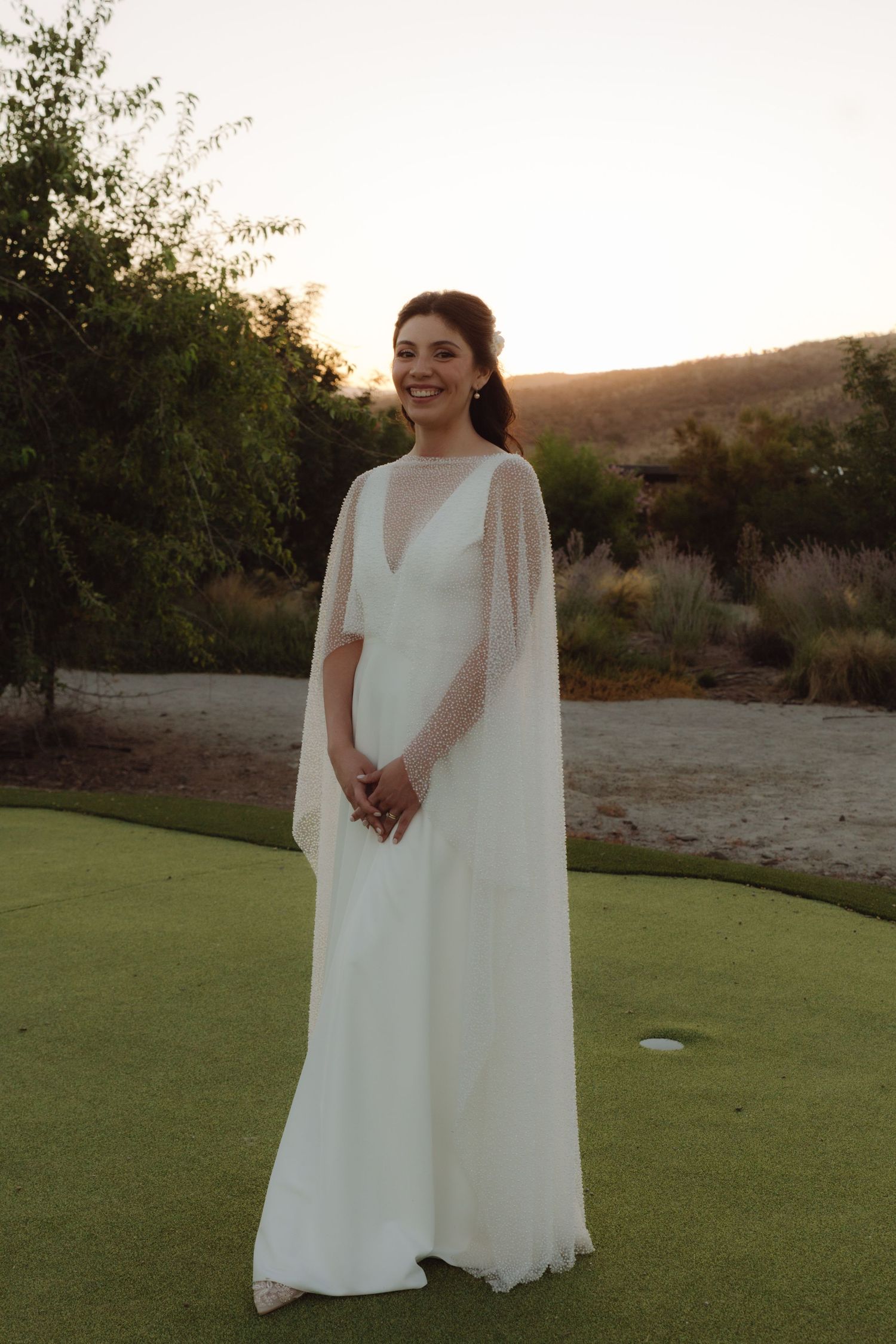 Smiling bride in elegant white cape wedding gown standing on green lawn at sunset.