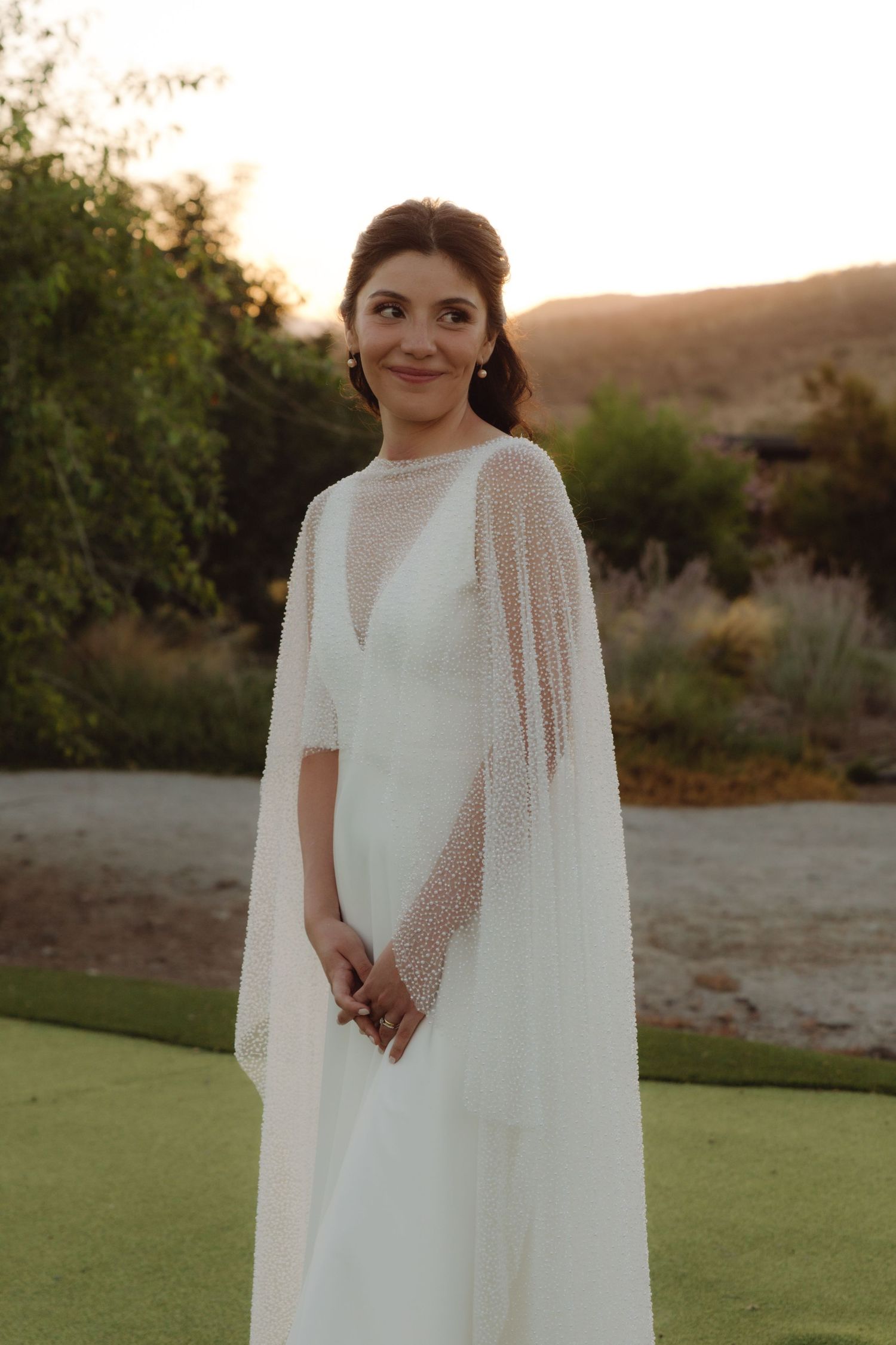 Bride in elegant white wedding gown with pearl beaded cape overlay posing outdoors at golden hour.