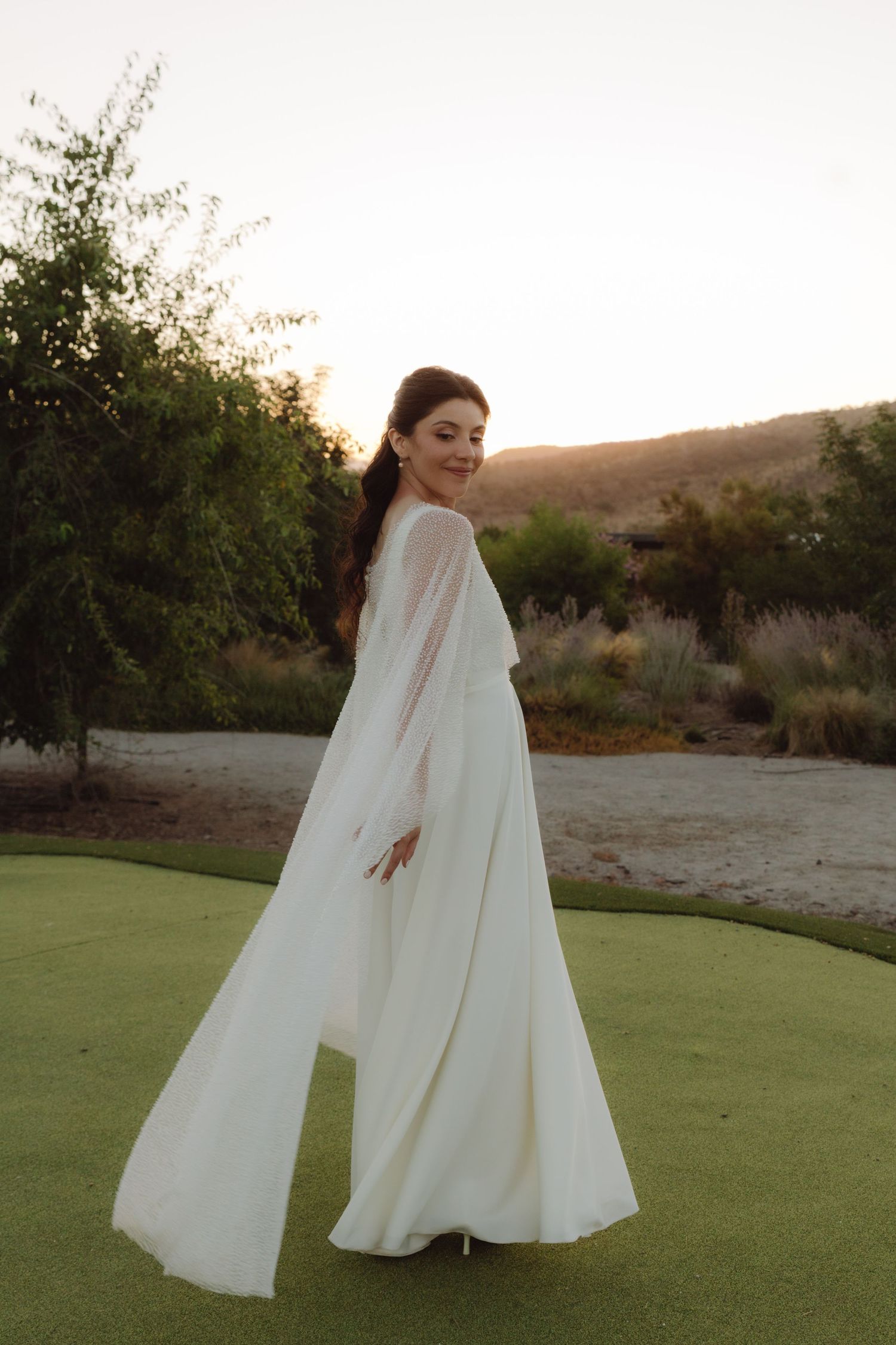 Bride twirling in flowing white wedding dress with sheer long sleeves on green lawn at sunset.