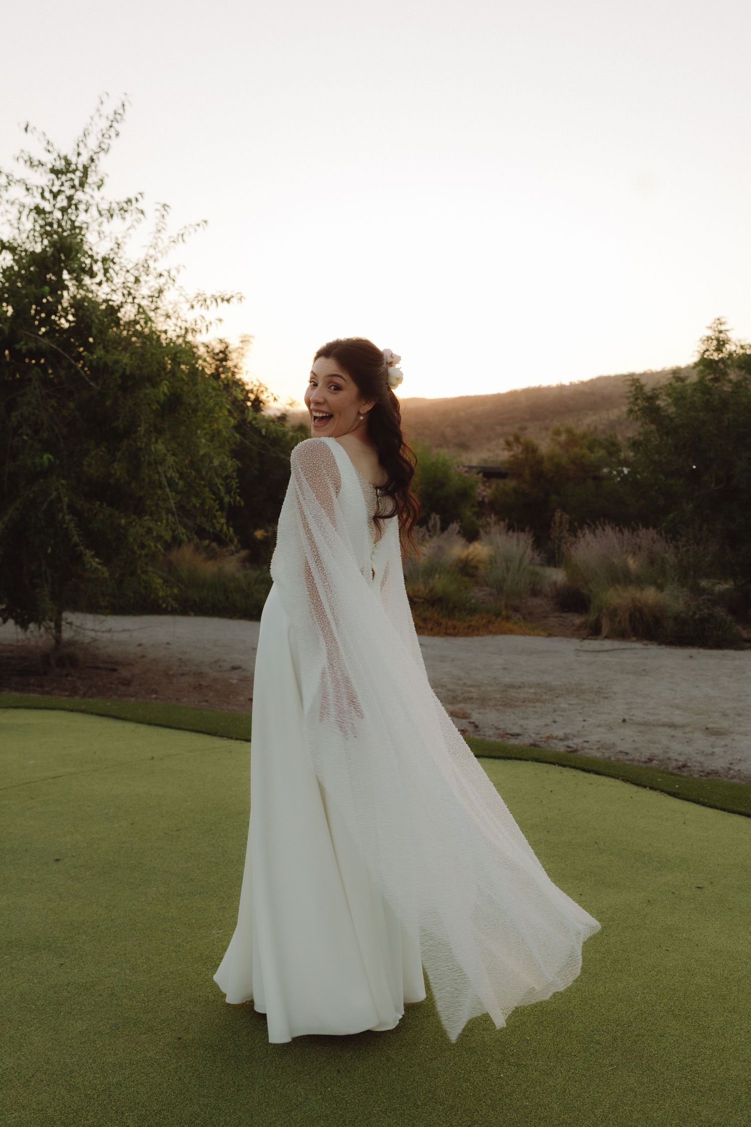 Joyful bride spinning in white chiffon wedding gown with flowing sleeves at outdoor garden venue.