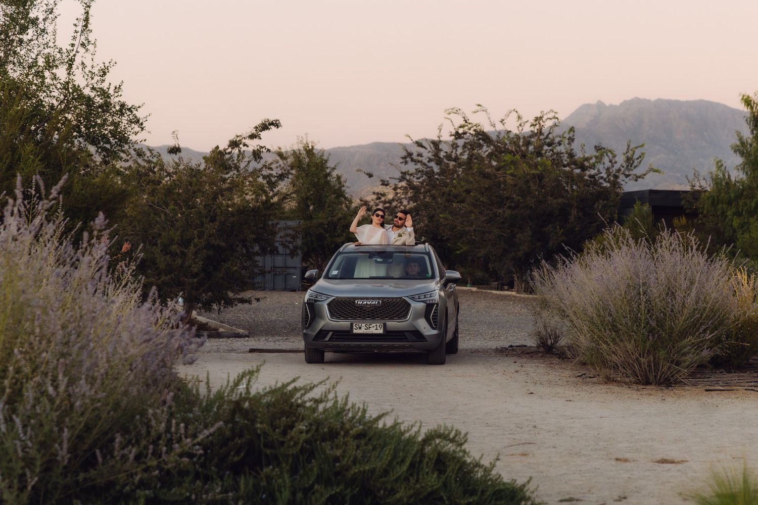 Gray SUV parked on gravel path surrounded by desert shrubs and mountains at dusk.