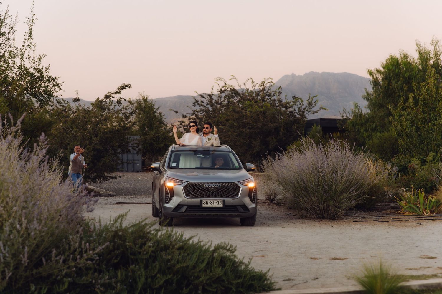 Gray SUV on gravel driveway with people posing on roof at desert wedding venue at sunset.