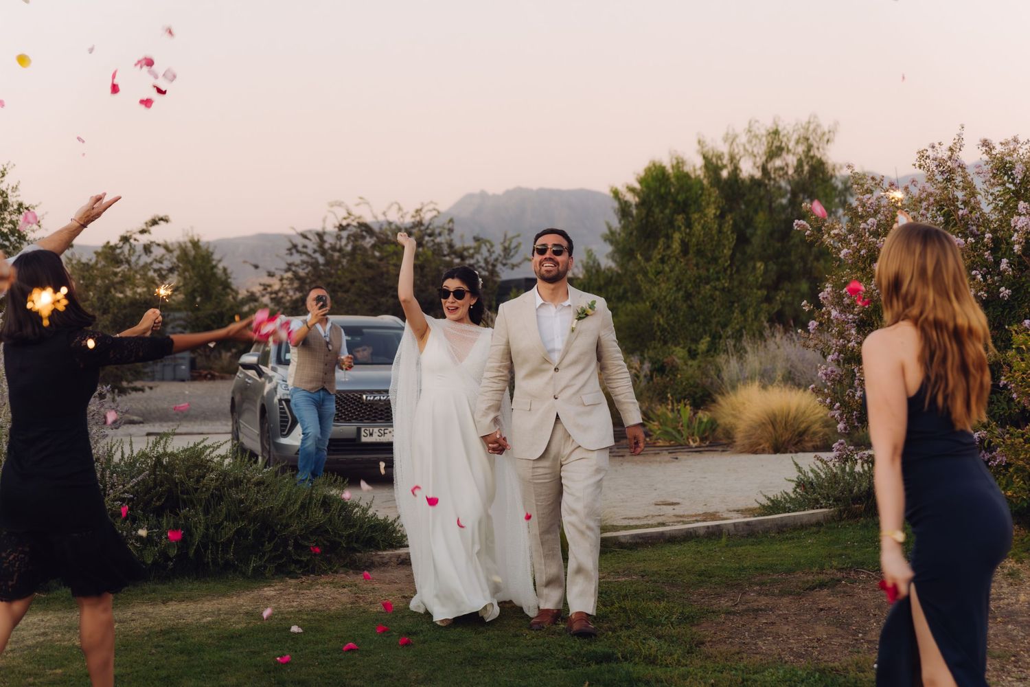 Newlyweds walking through rose petal toss by guests at outdoor wedding celebration at dusk.