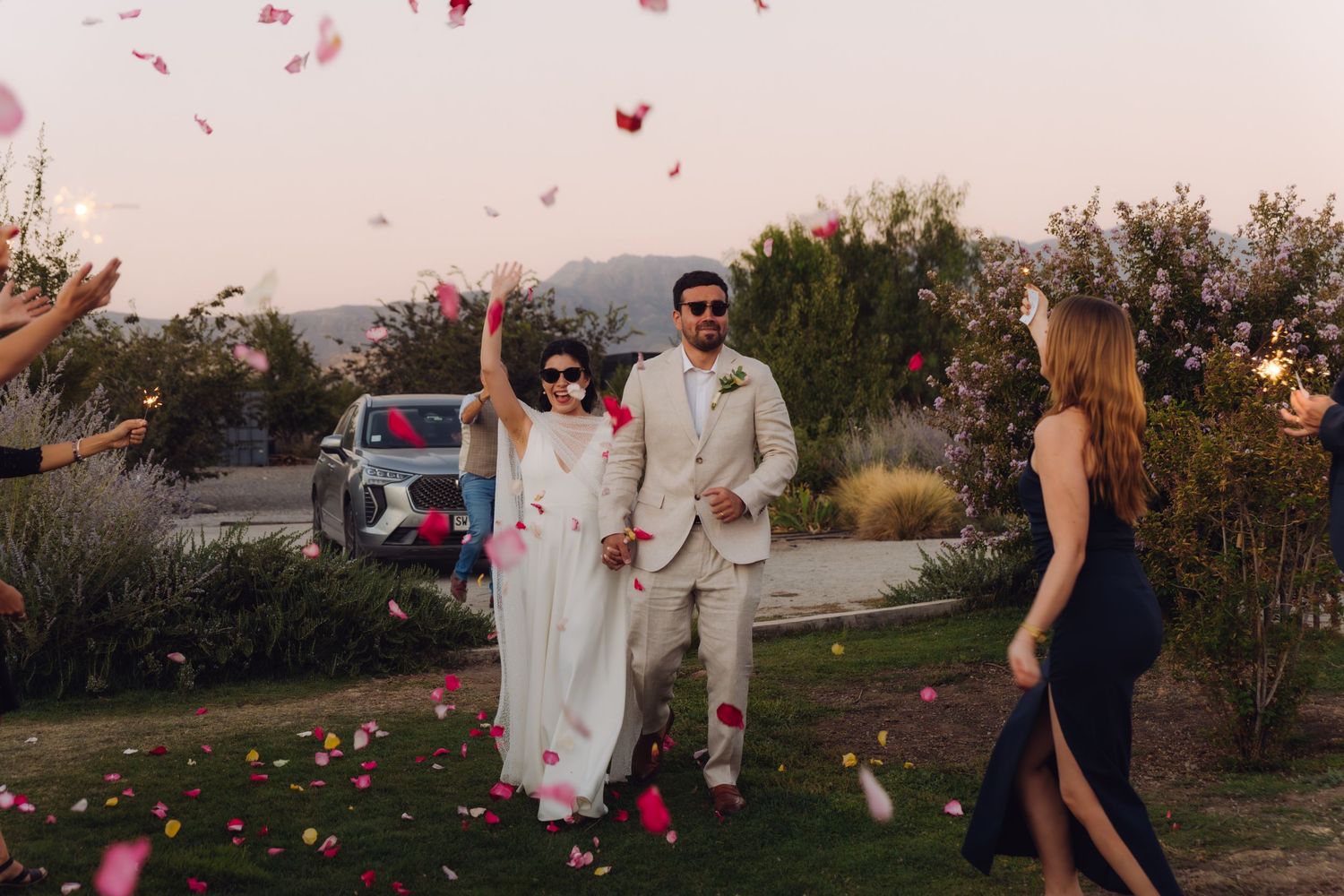 Happy couple walking through festive rose petal confetti toss at outdoor wedding reception exit.