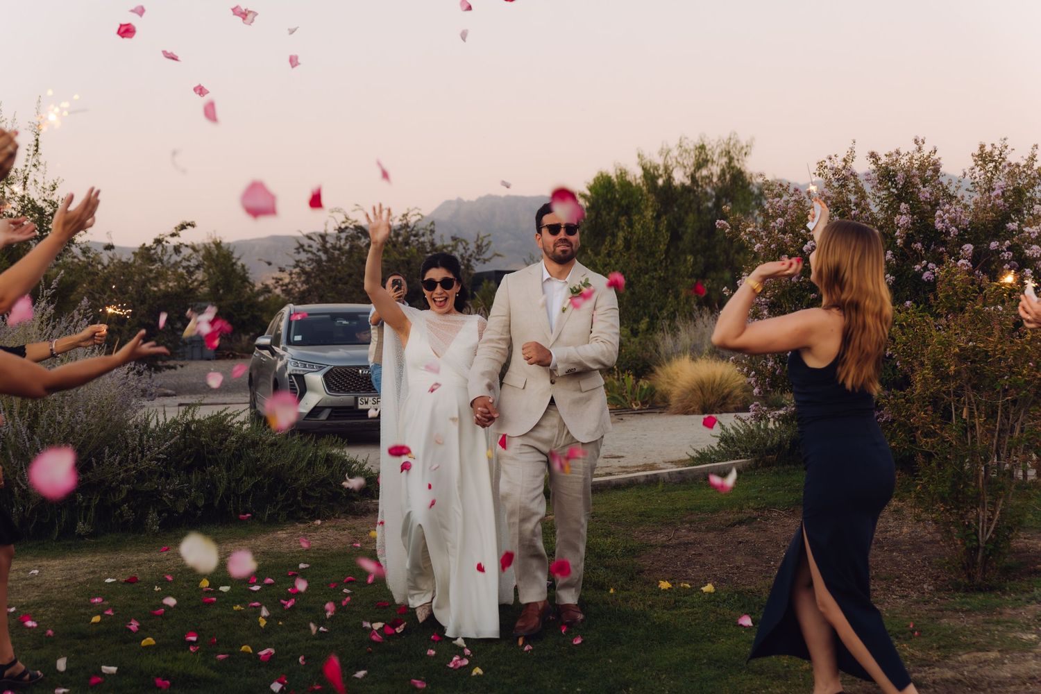 Bride and groom celebrating with rose petal toss by guests at outdoor wedding at dusk.
