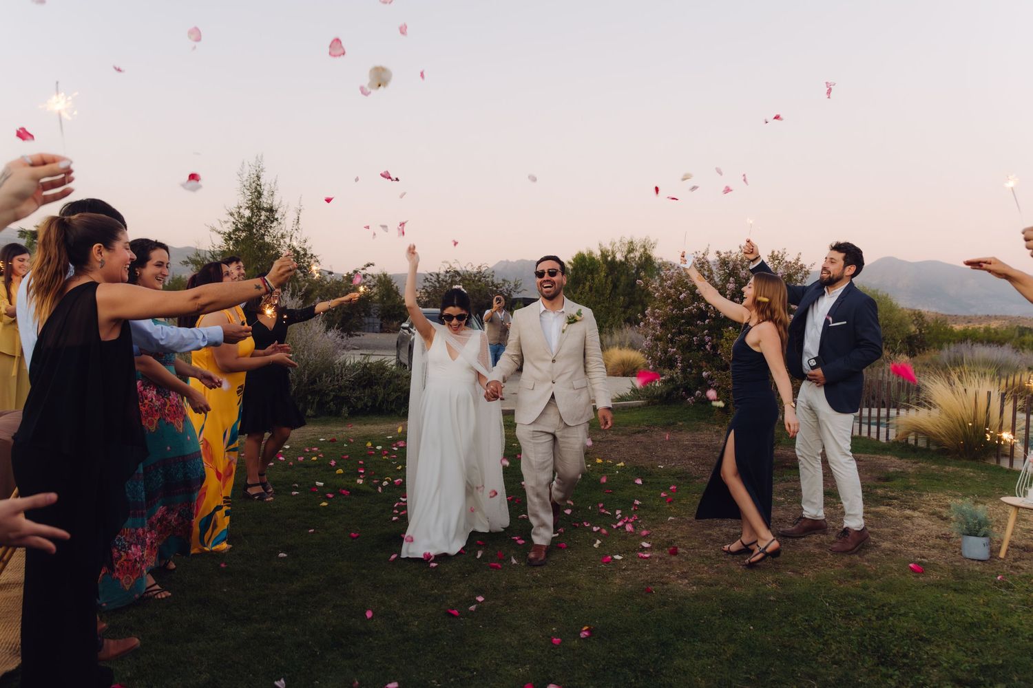 Bride and groom walk through cheering guests tossing rose petals at outdoor wedding celebration.