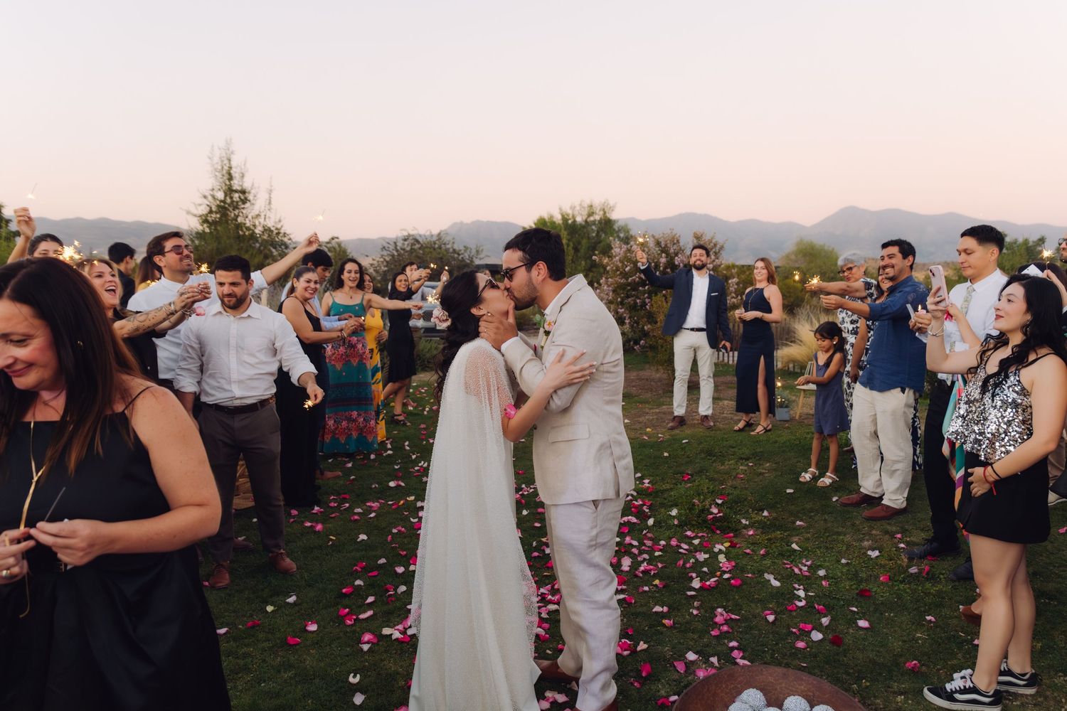 Newlyweds share romantic kiss surrounded by applauding guests at outdoor garden wedding ceremony.