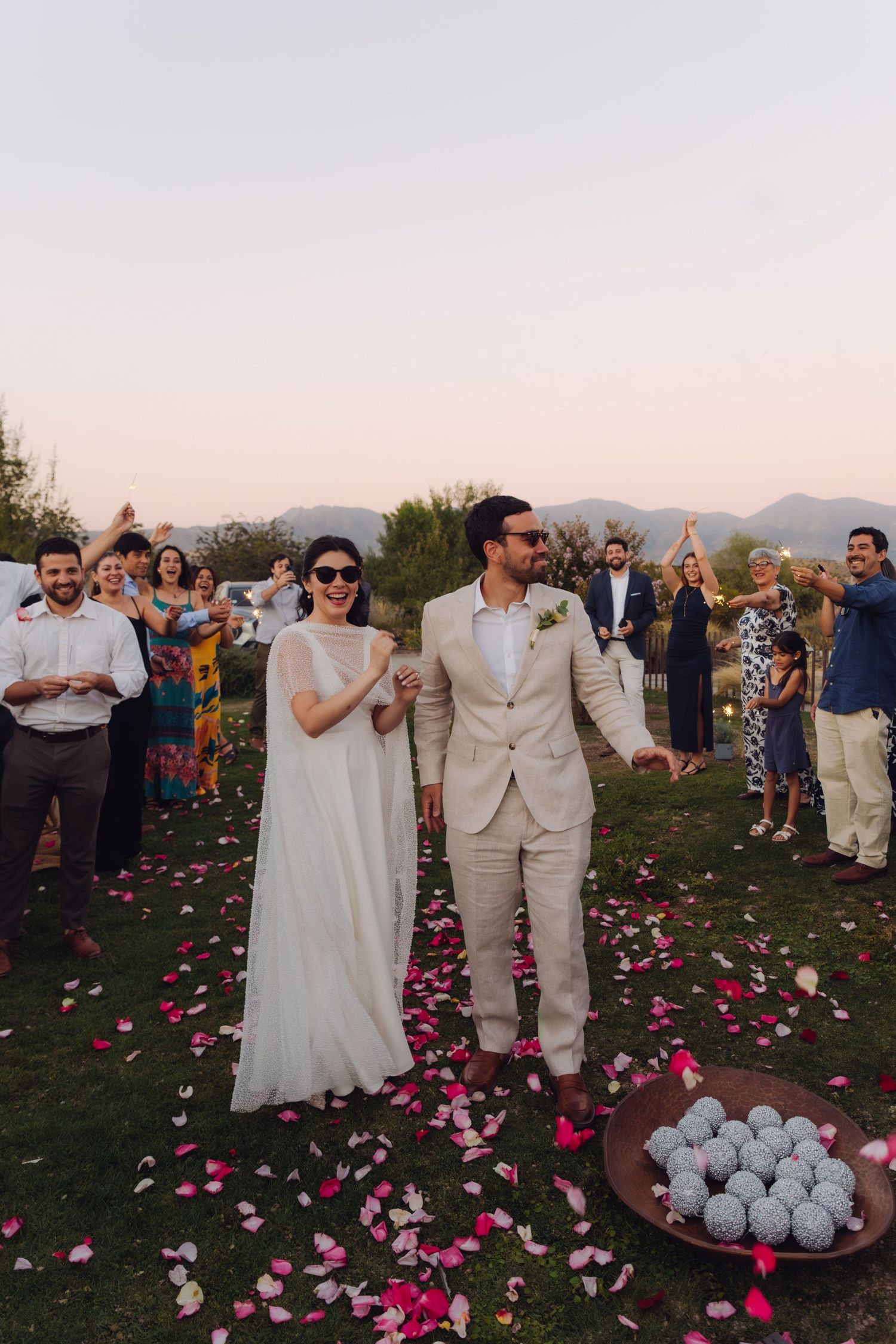Smiling newlyweds walk rose petal lined path as guests celebrate at outdoor mountain wedding.