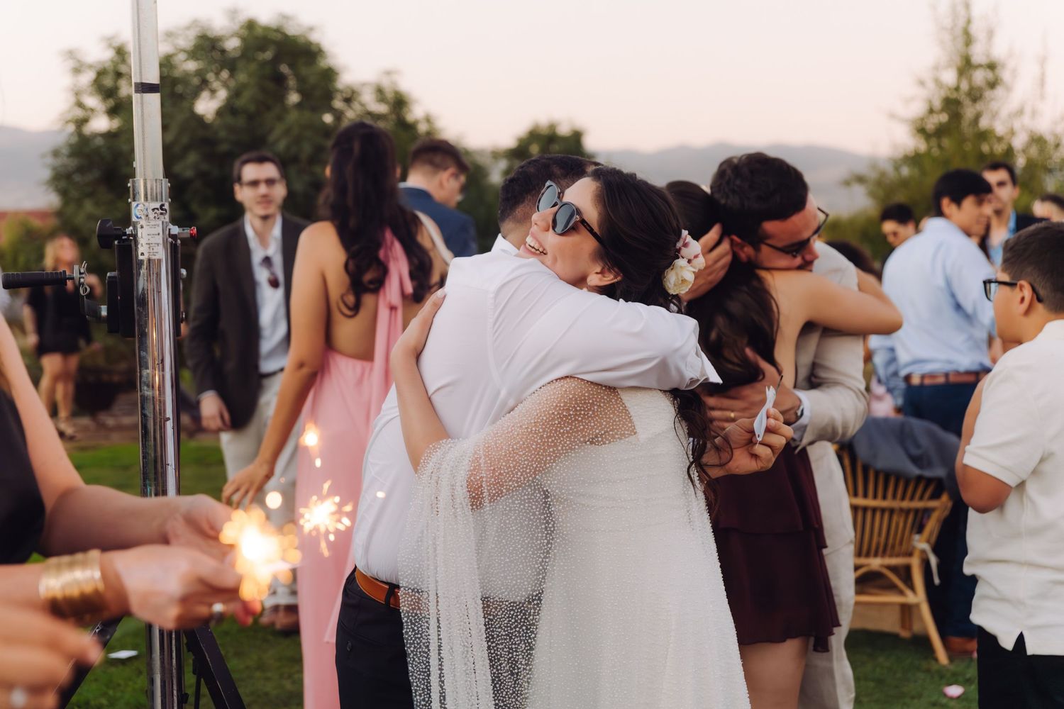 Bride in white gown embraces wedding guest while others hold sparklers at evening celebration.