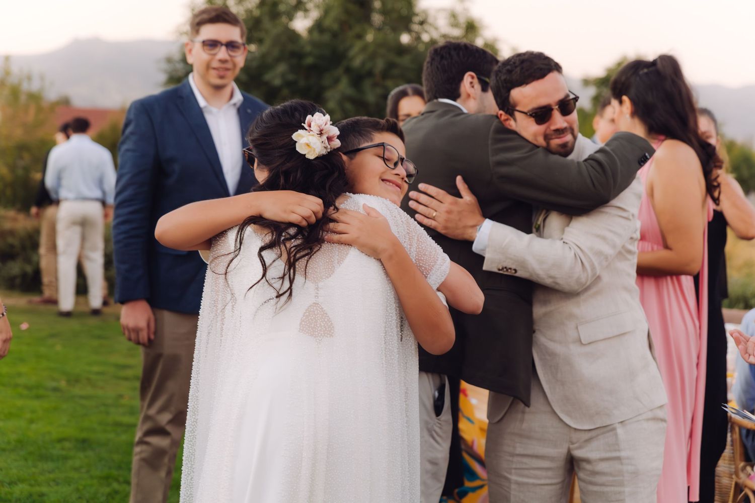 Groom in beige suit hugs young flower girl with floral crown at outdoor wedding celebration.