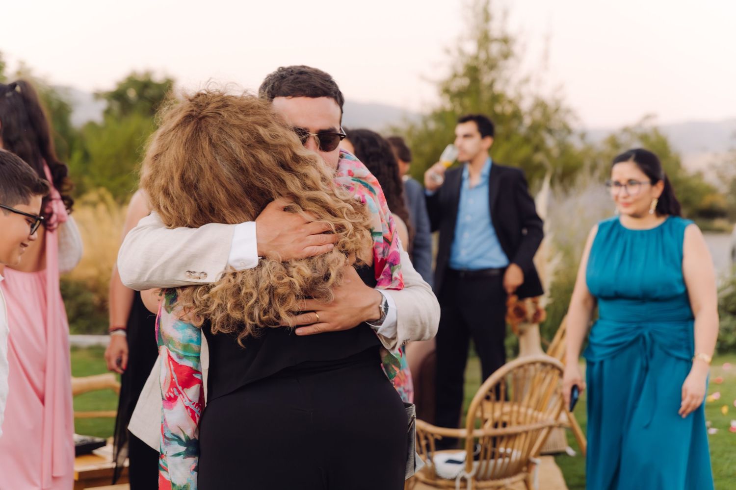 Groom in sunglasses shares heartfelt hug with wedding guest at outdoor garden reception.