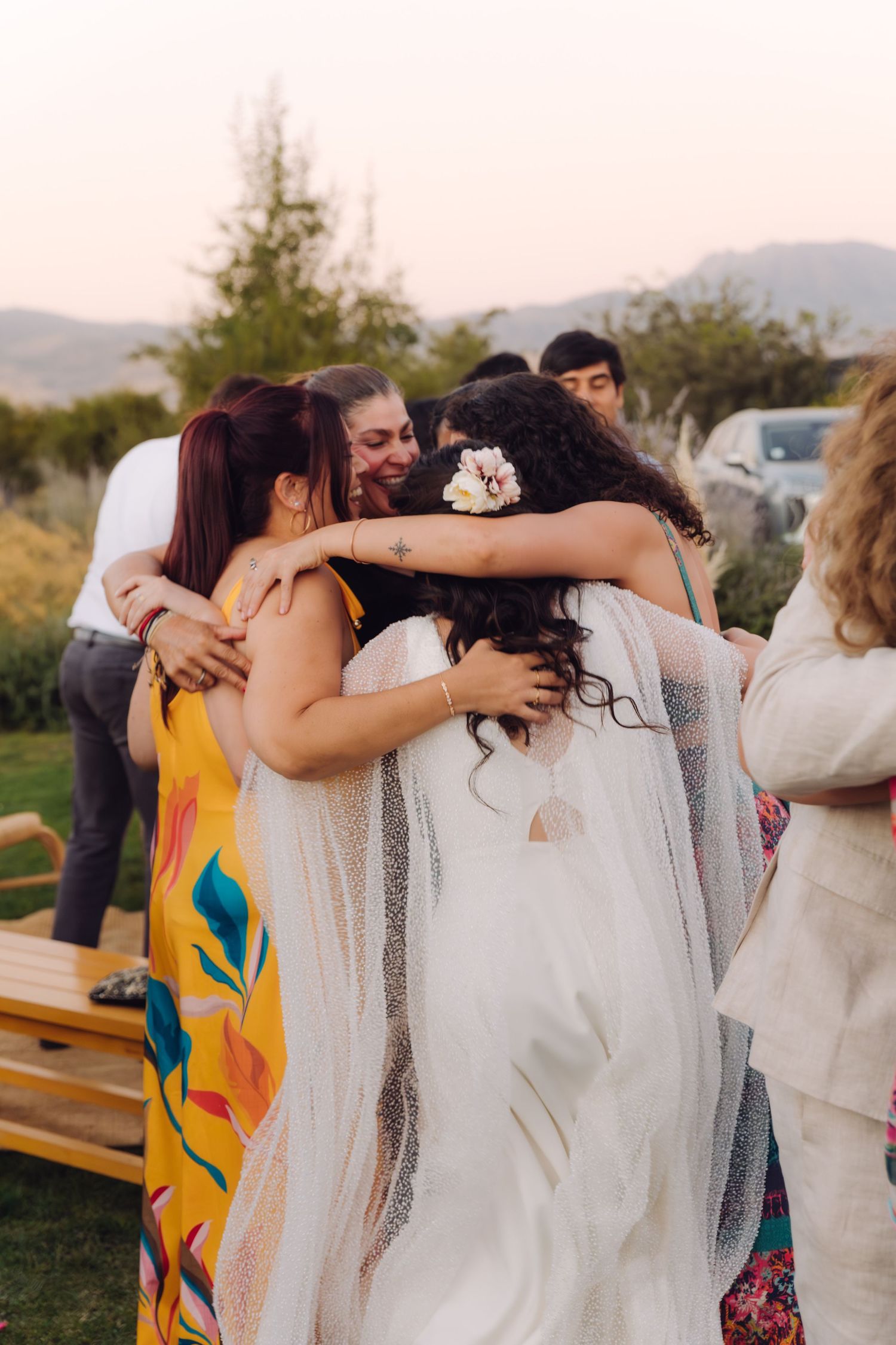 Bride with floral hair accessory shares group hug with two guests in colorful dresses outdoors.