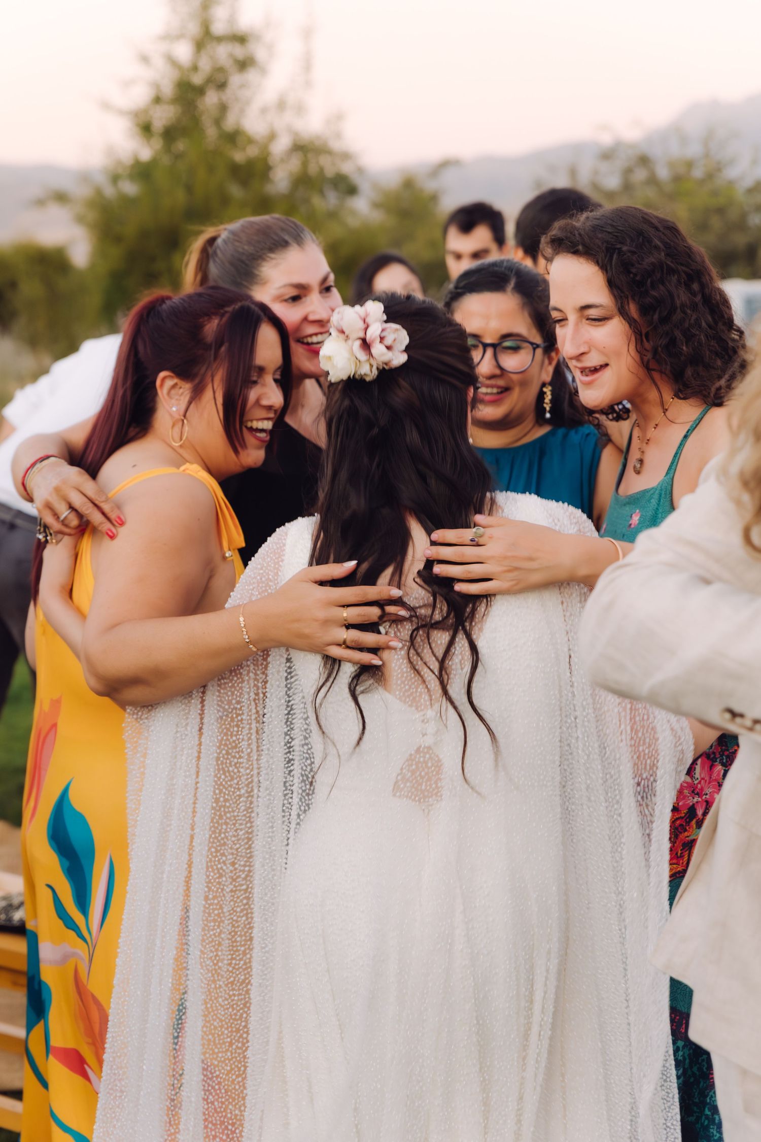 Bride surrounded by joyful female guests in a warm group hug at an outdoor wedding celebration.