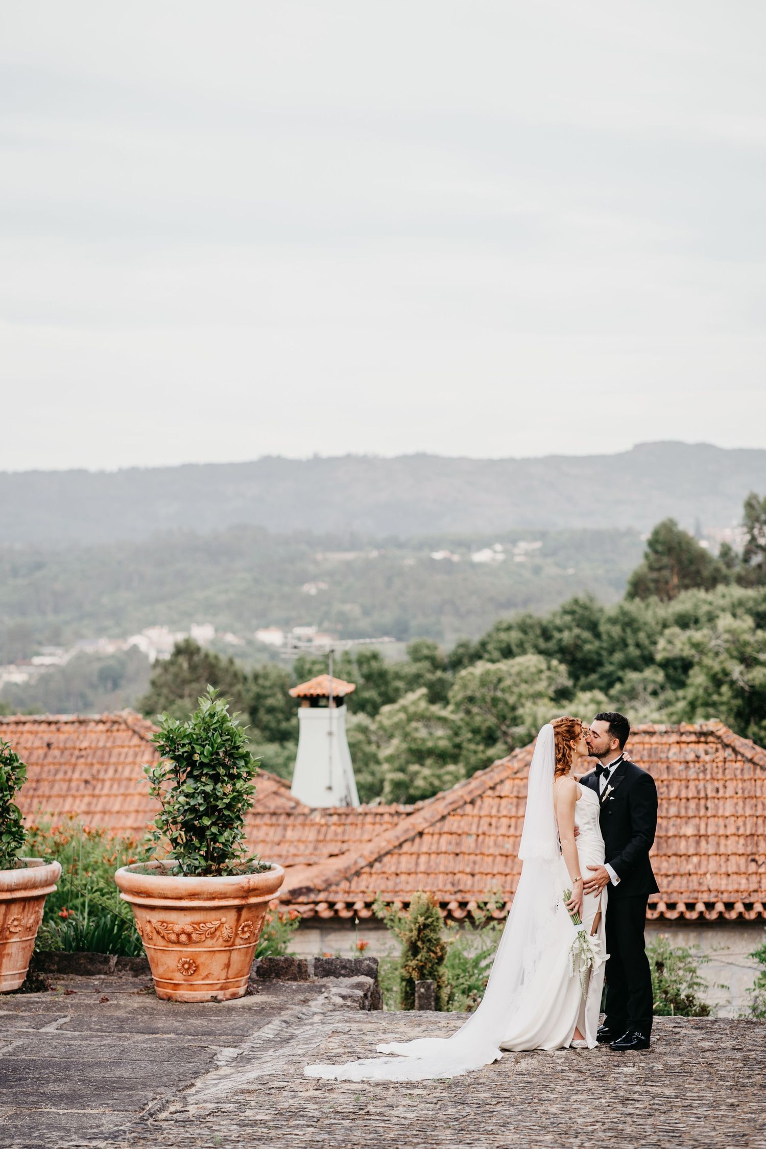 Bride and groom sharing a kiss on a terrace with terracotta rooftops and lush green hills in the background.
