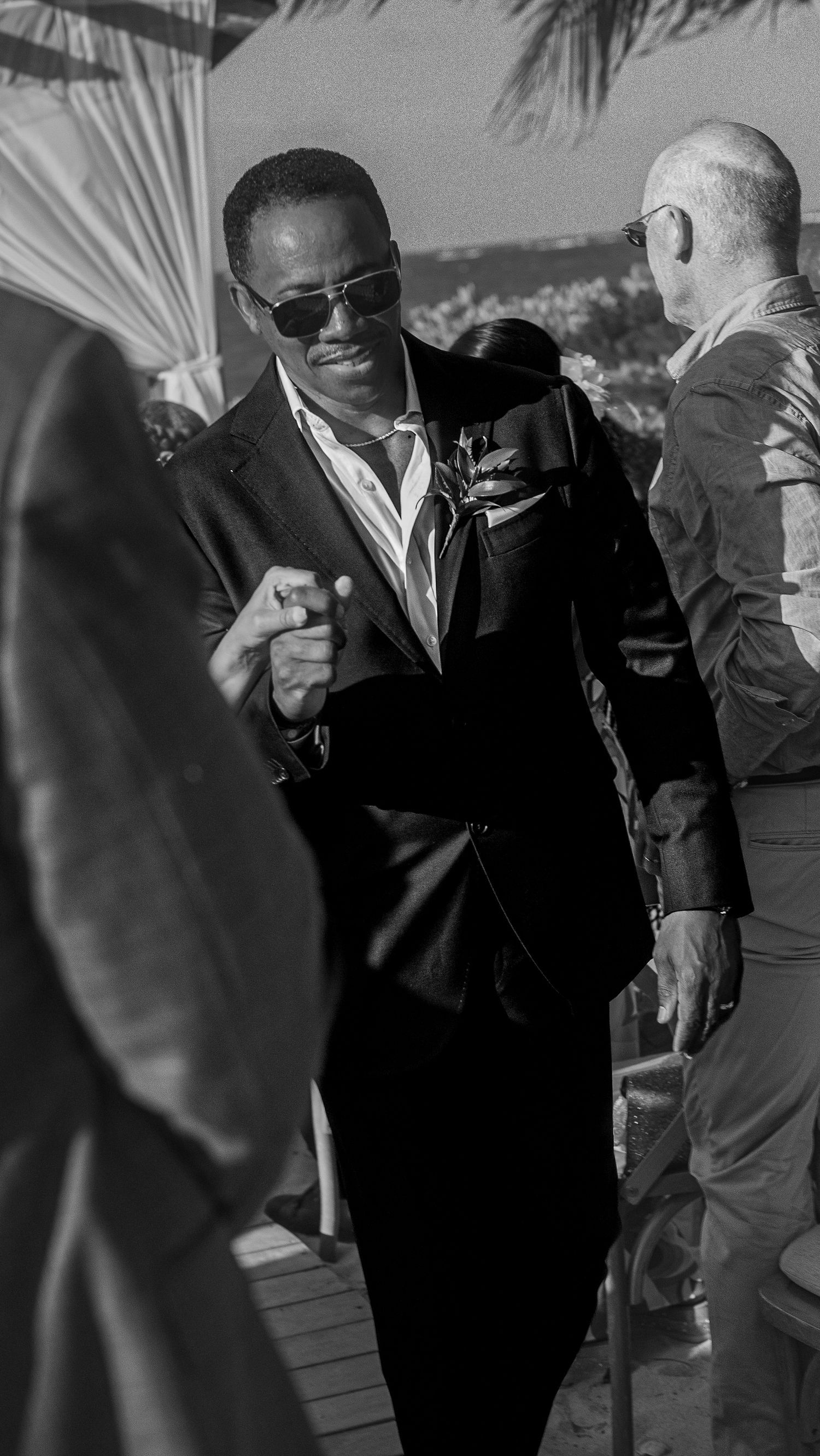 Black and white photo of a well-dressed man in sunglasses and suit at an outdoor wedding event.