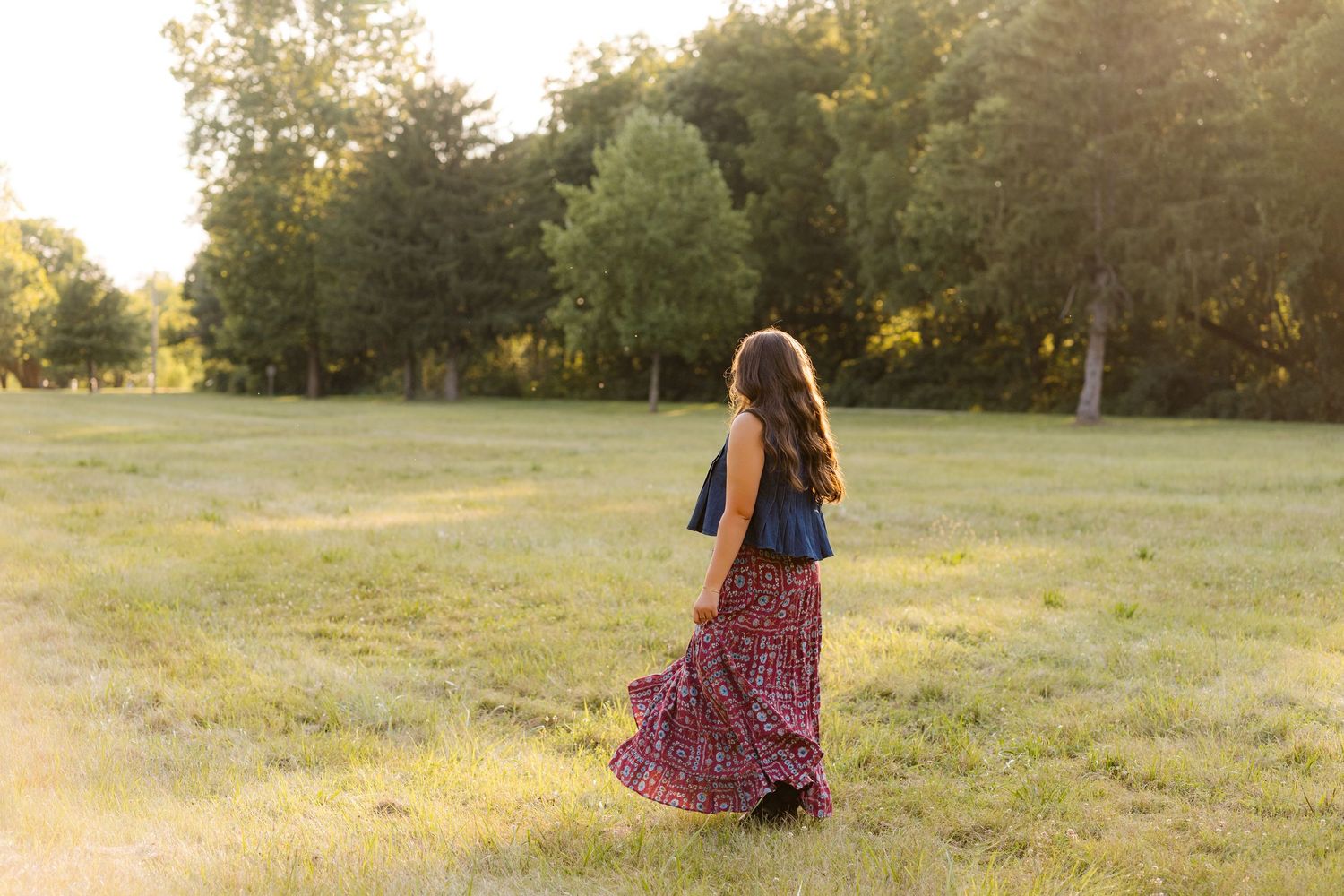 Young woman in floral maxi skirt walks through sunlit green meadow surrounded by trees during golden hour.