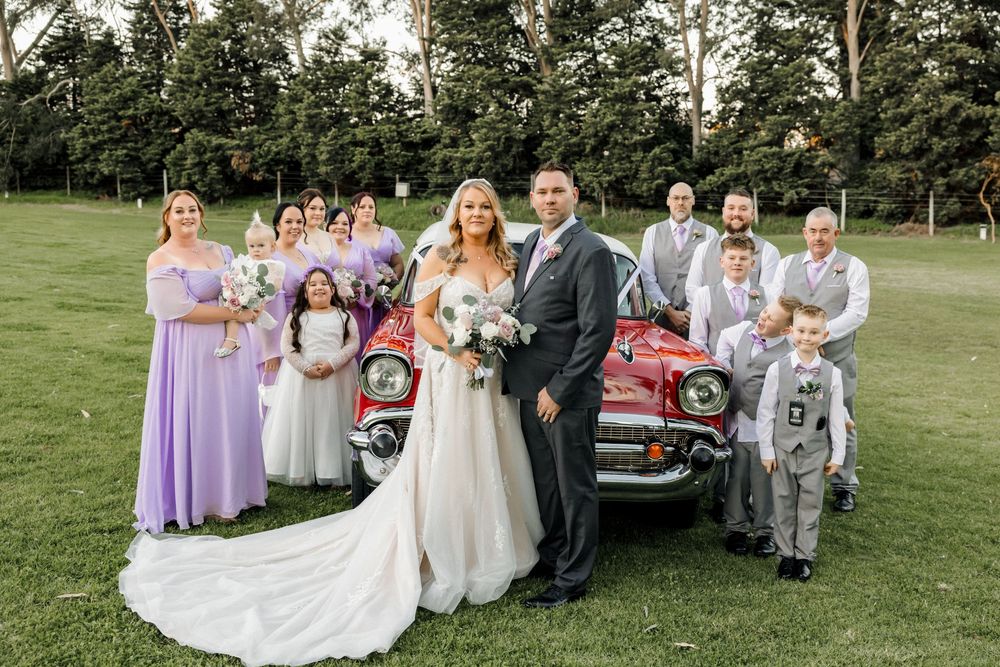 Wedding party poses with classic red car on green lawn, bride and groom centered with bridesmaids in lavender and groomsmen.