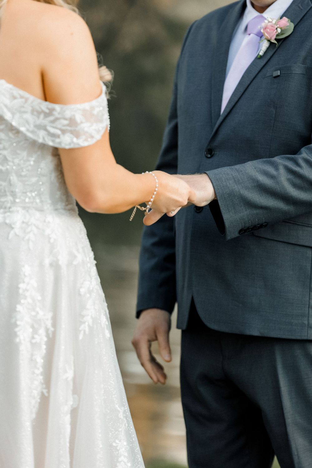 Bride and groom holding hands during wedding ceremony, bride in lace off-shoulder gown, groom in navy suit with floral boutonniere.
