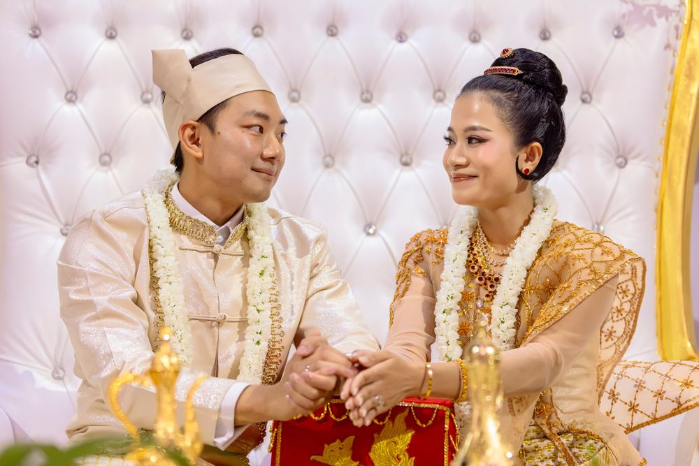 Couple in traditional Southeast Asian wedding attire holding hands, bride in gold dress, groom in white ceremonial outfit.
