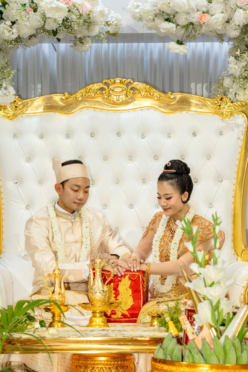Bride and groom in traditional Southeast Asian wedding attire seated on ornate gold throne during ceremony.
