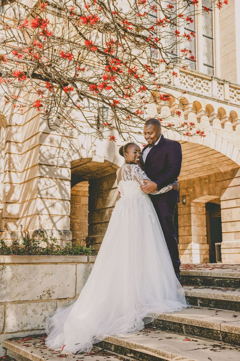 Elegant Black couple in wedding attire embracing on stone steps beneath a red flowering tree.
