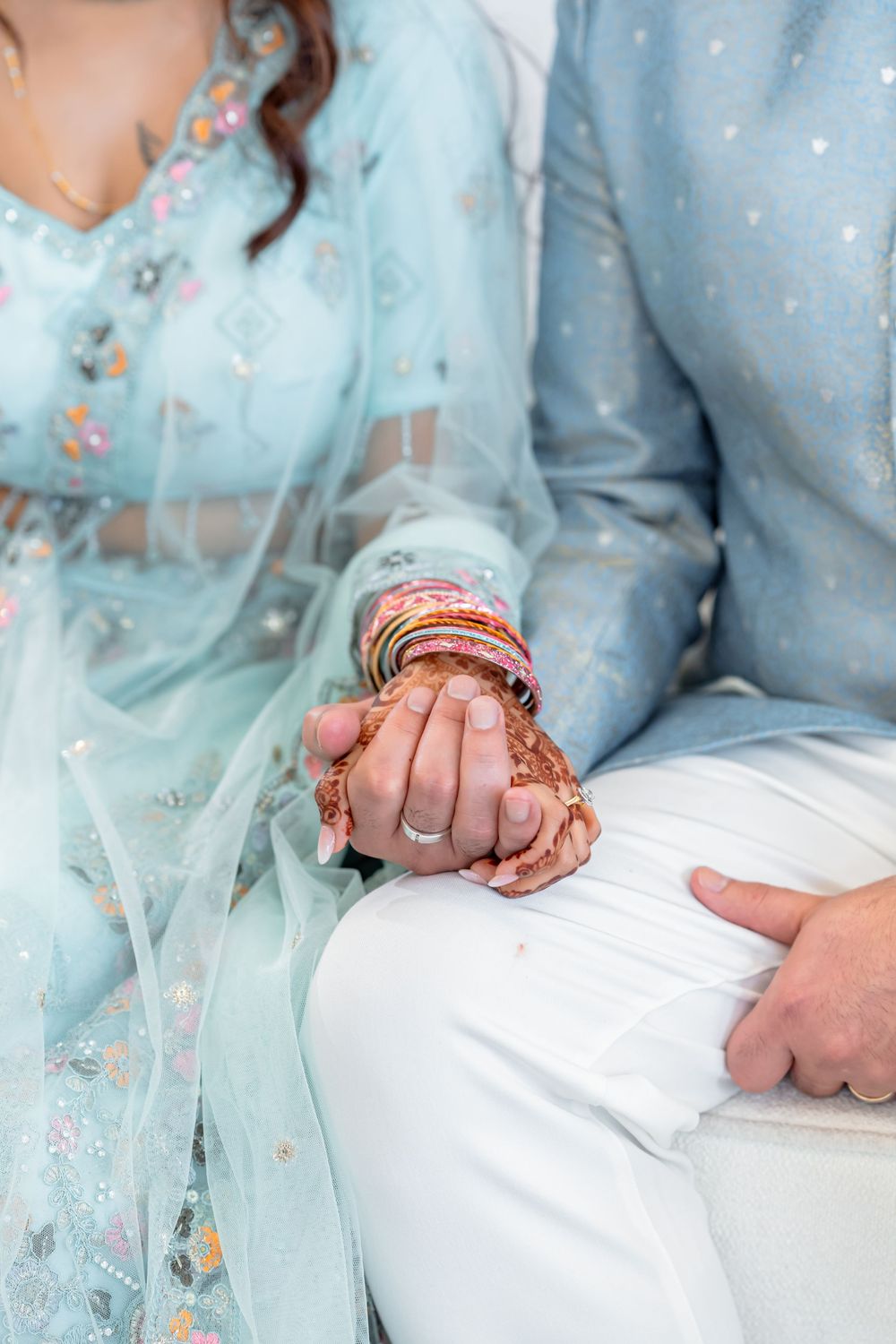 Indian bride with mehndi and bangles holds groom's hand during wedding ceremony, both wearing light blue outfits.