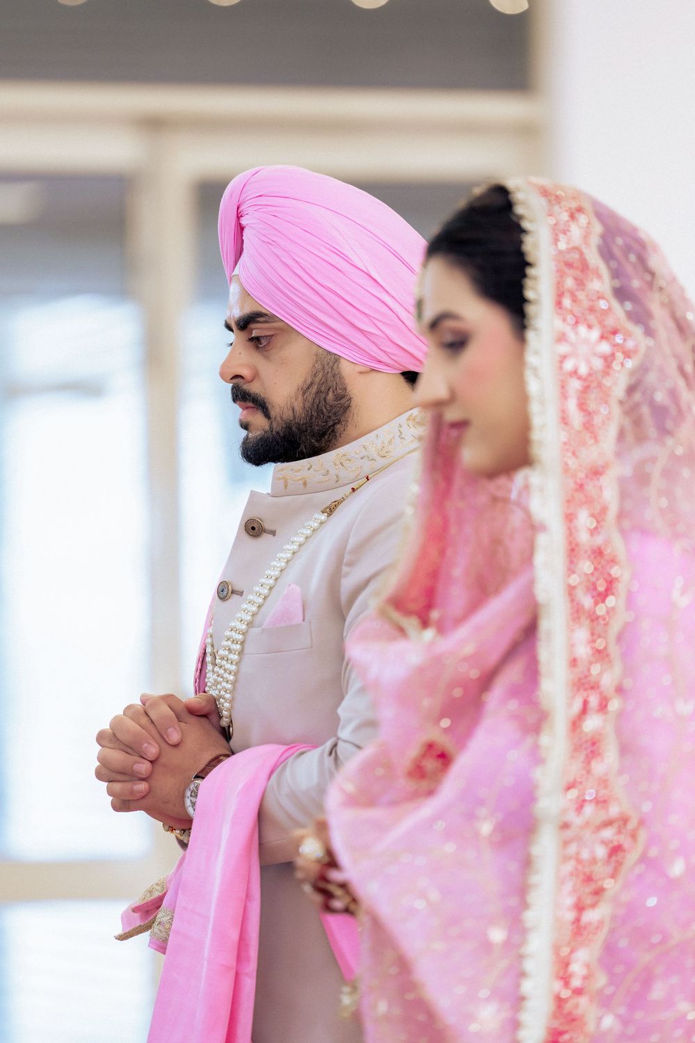 Sikh couple in traditional wedding attire, groom in pink turban and bride in embroidered pink dupatta, praying together.