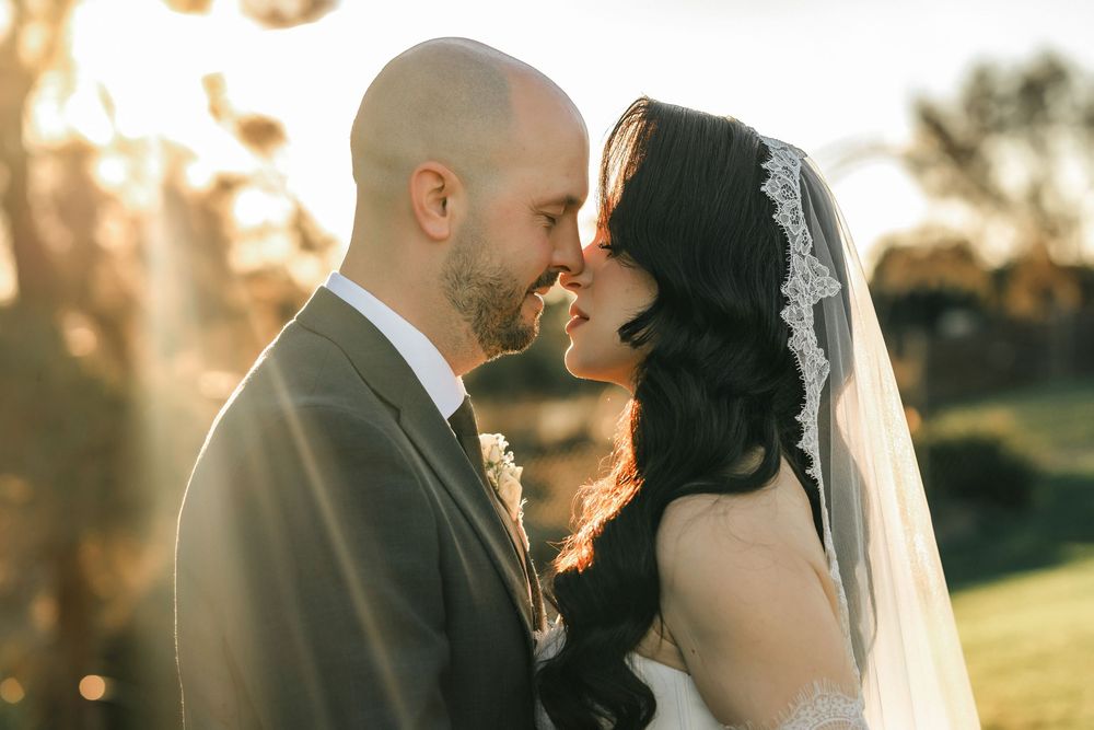 A bride and groom share a romantic forehead touch at golden hour, with the bride wearing a lace veil and flowing dark hair.