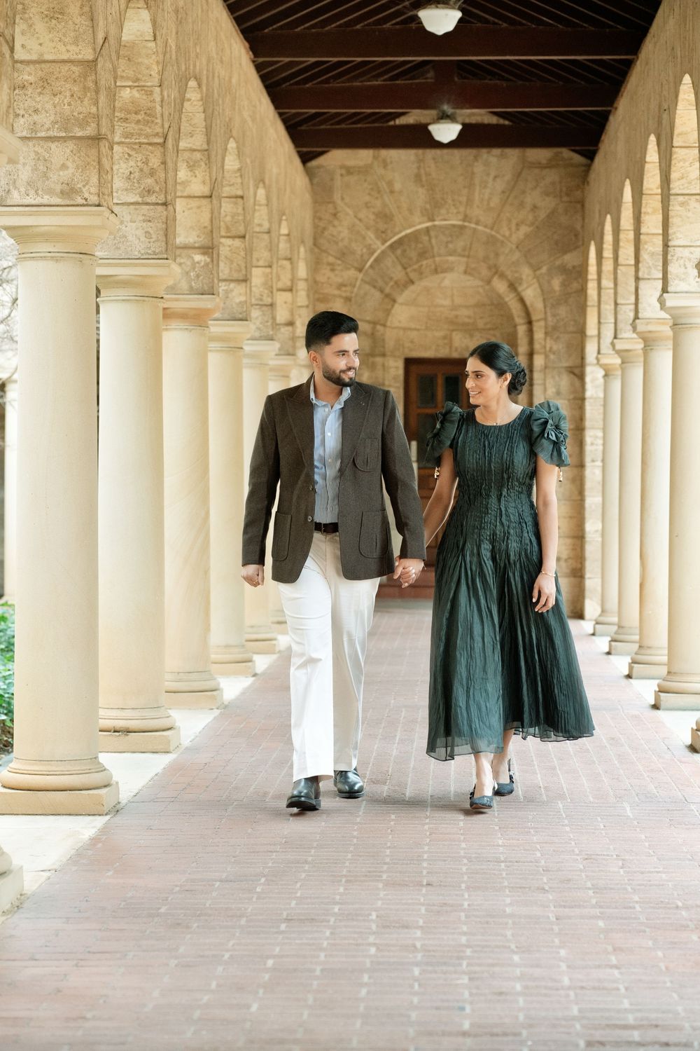 Couple walking hand-in-hand through elegant stone archway colonnade, man in blazer and woman in green dress.