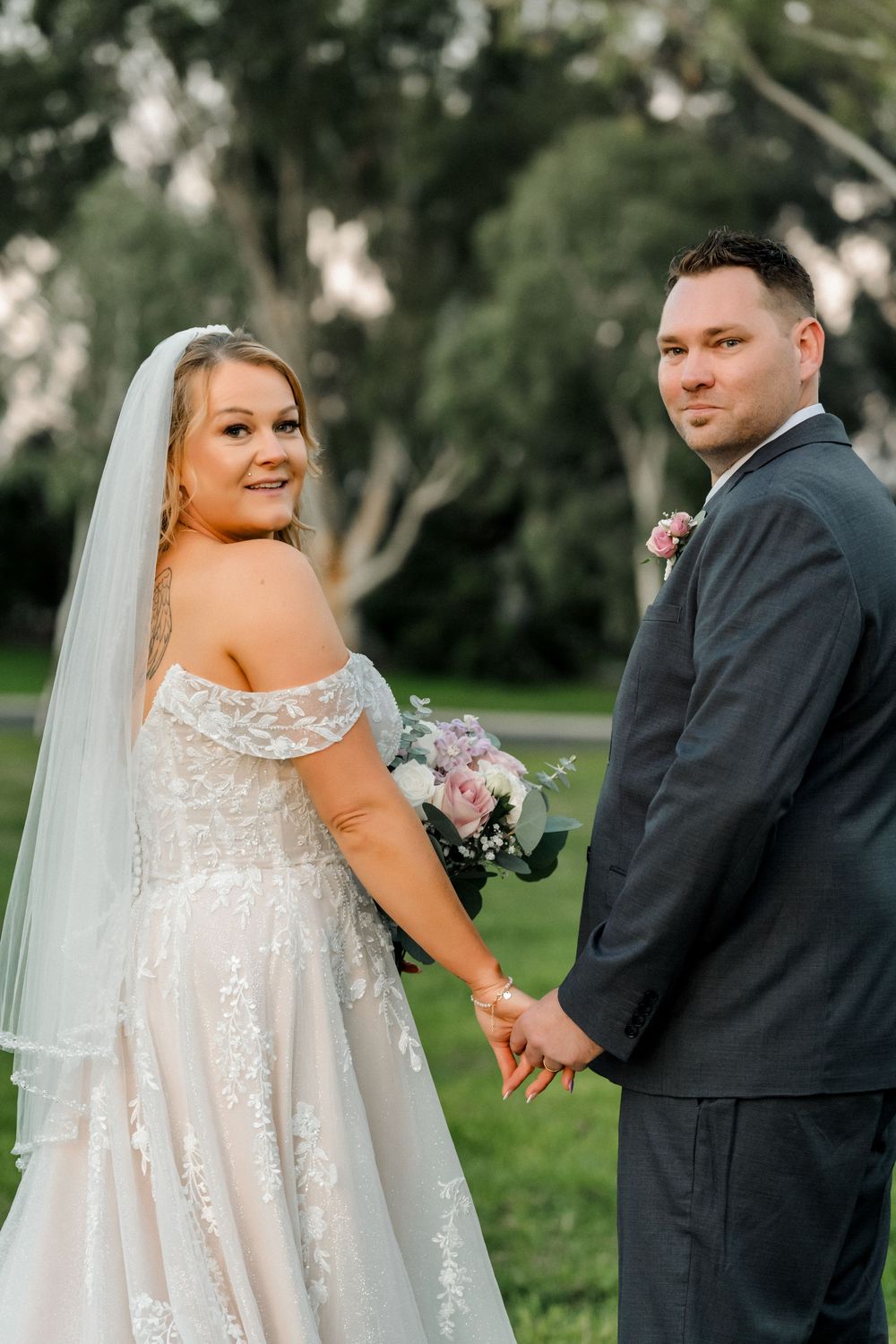 Bride in lace off-shoulder gown and veil holds hands with groom in dark suit, posing outdoors among green trees.