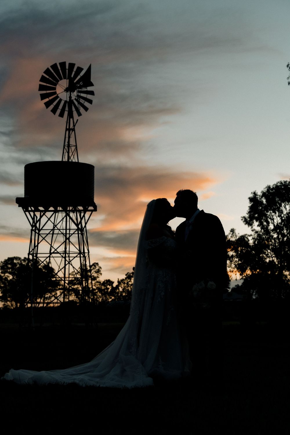 Silhouette of a bride and groom kissing at sunset beside a rustic windmill and water tower.