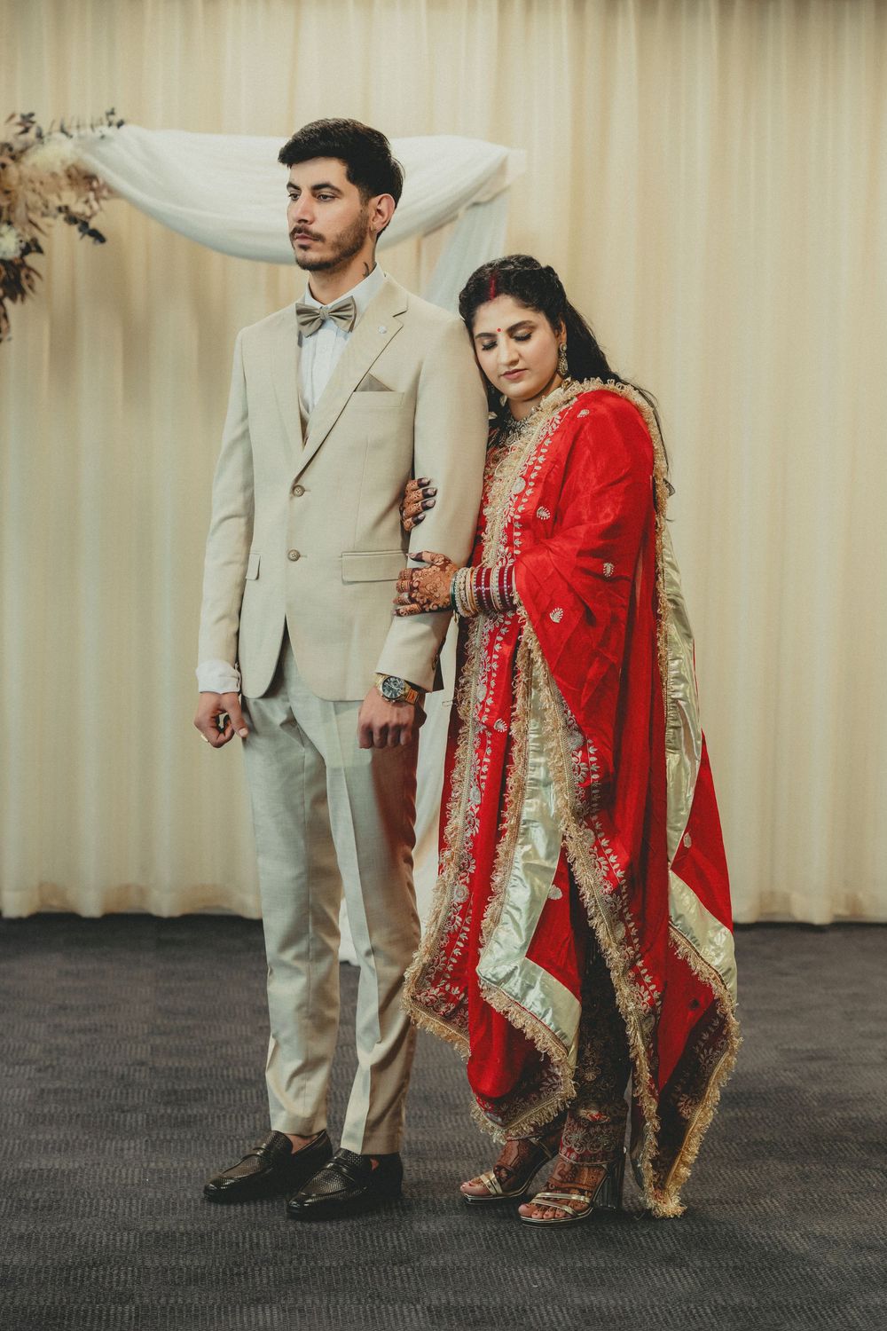 Bride in red embroidered lehenga leans on groom in beige suit at South Asian wedding ceremony.