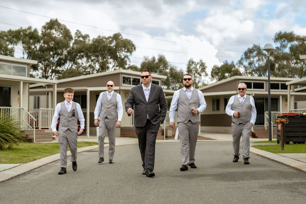 Groom and groomsmen in grey suits walking confidently outdoors at a wedding venue on a sunny day.