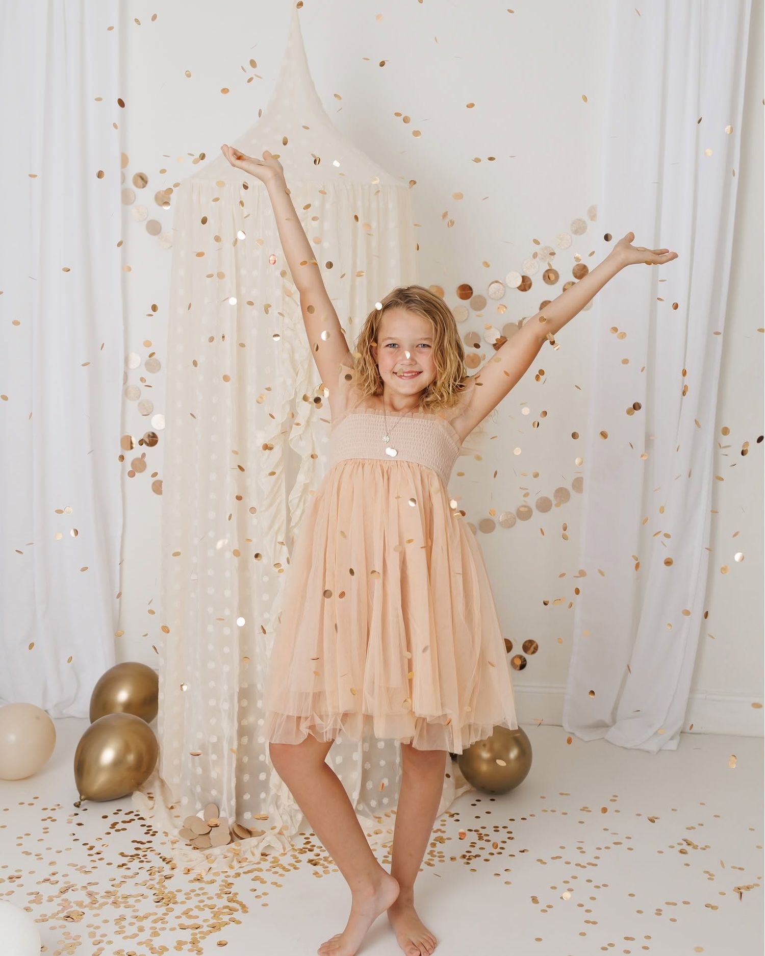 A joyful girl in a peach tulle dress celebrates with gold confetti falling around her, arms raised, surrounded by gold balloons.