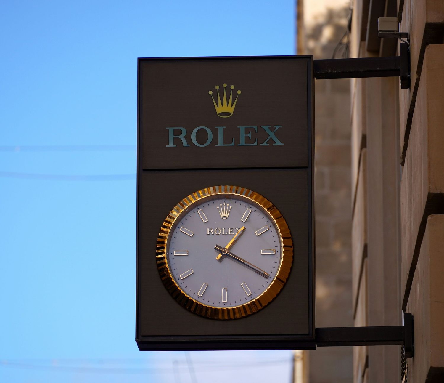 Rolex branded outdoor clock sign with gold trim mounted on a building exterior against a clear blue sky.