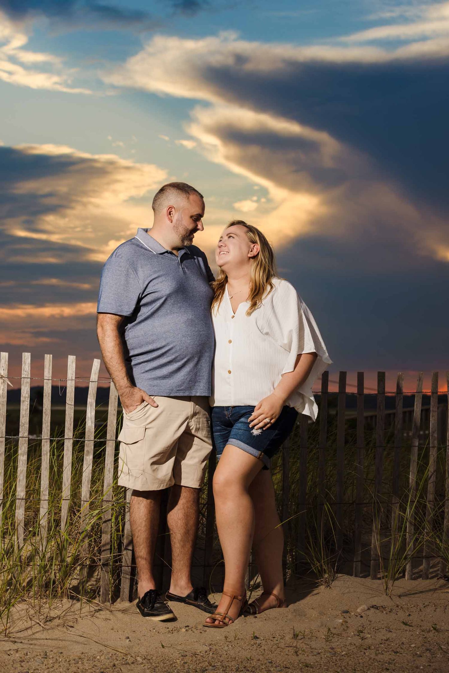 Couple sharing a romantic moment on a sandy beach at sunset with dramatic clouds and a white fence backdrop.