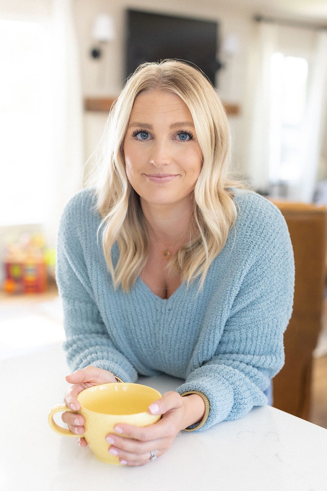Smiling blonde woman in light blue sweater and jeans sitting relaxed on brown leather sofa at home.