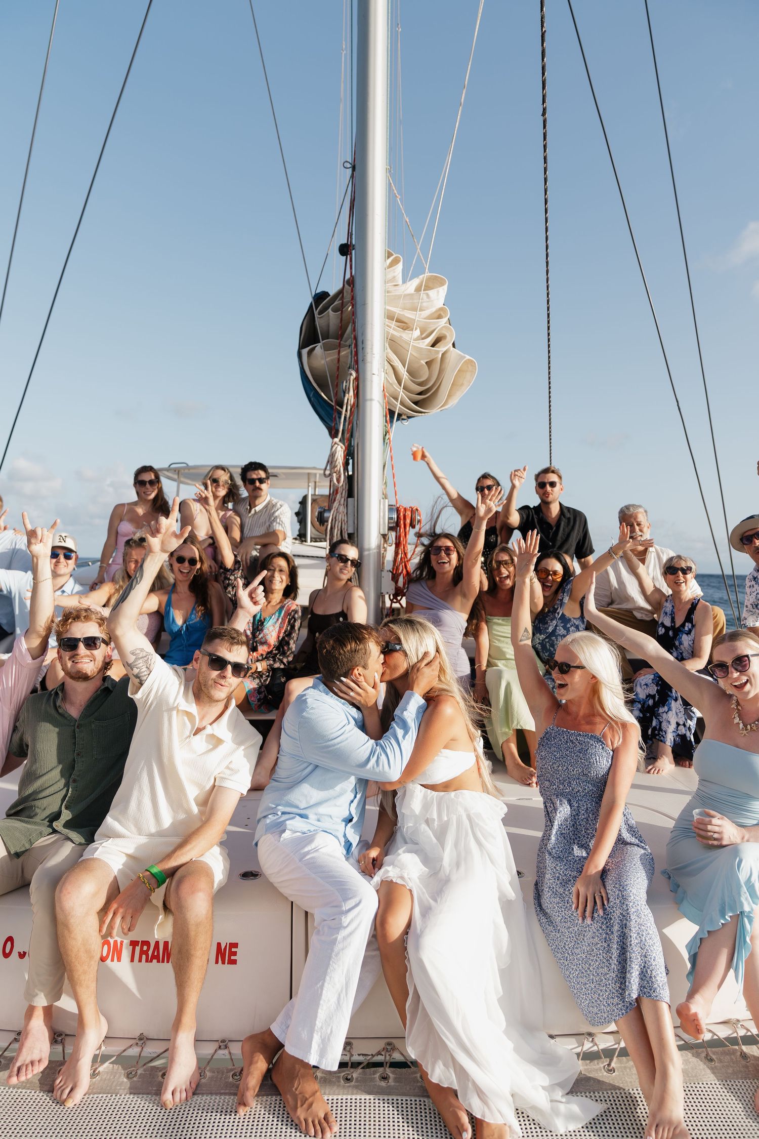 Couple kissing on a sailboat surrounded by cheering guests during a sunny destination wedding celebration.