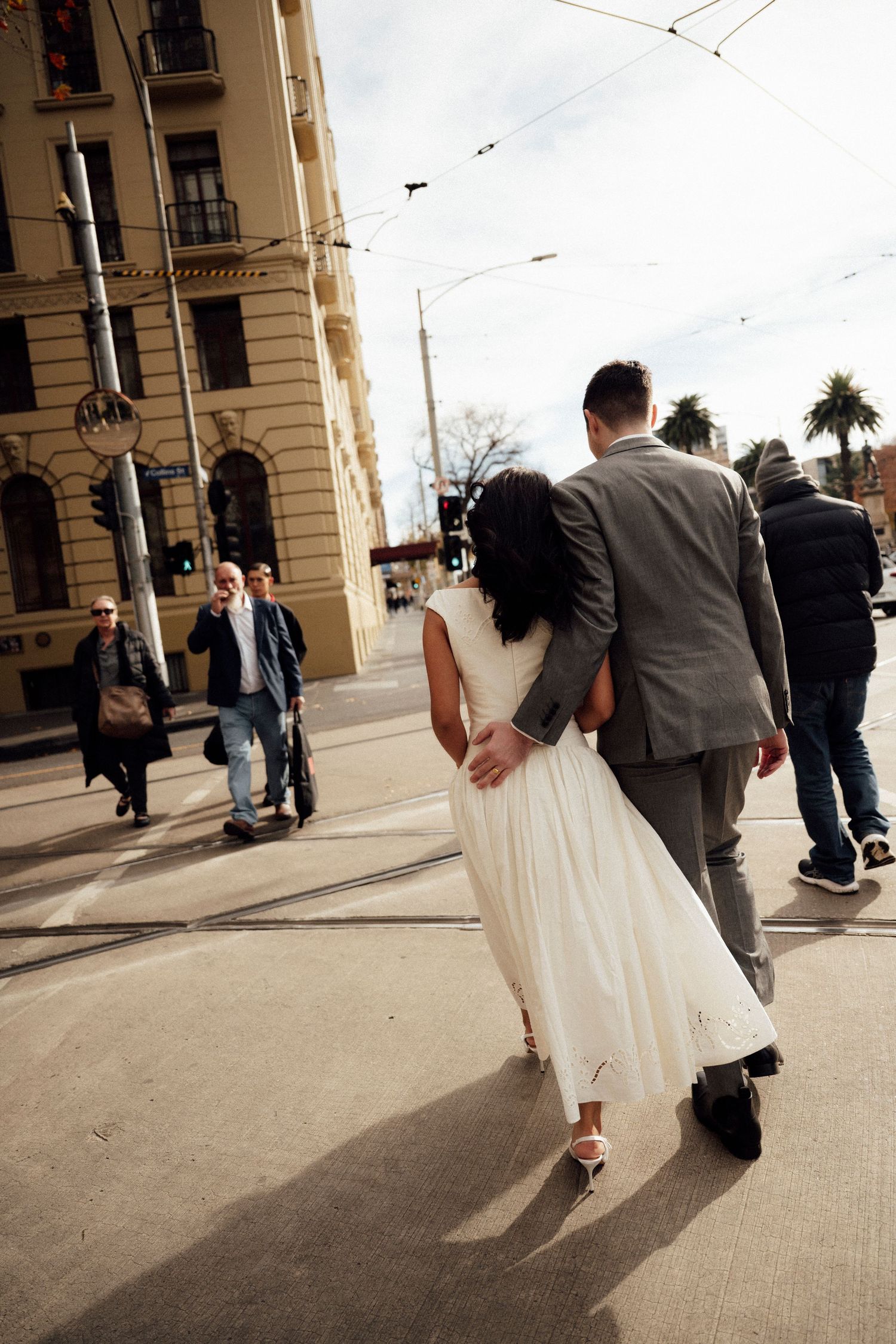Bride and groom walking hand in hand across a city street intersection, with pedestrians and historic buildings in background.