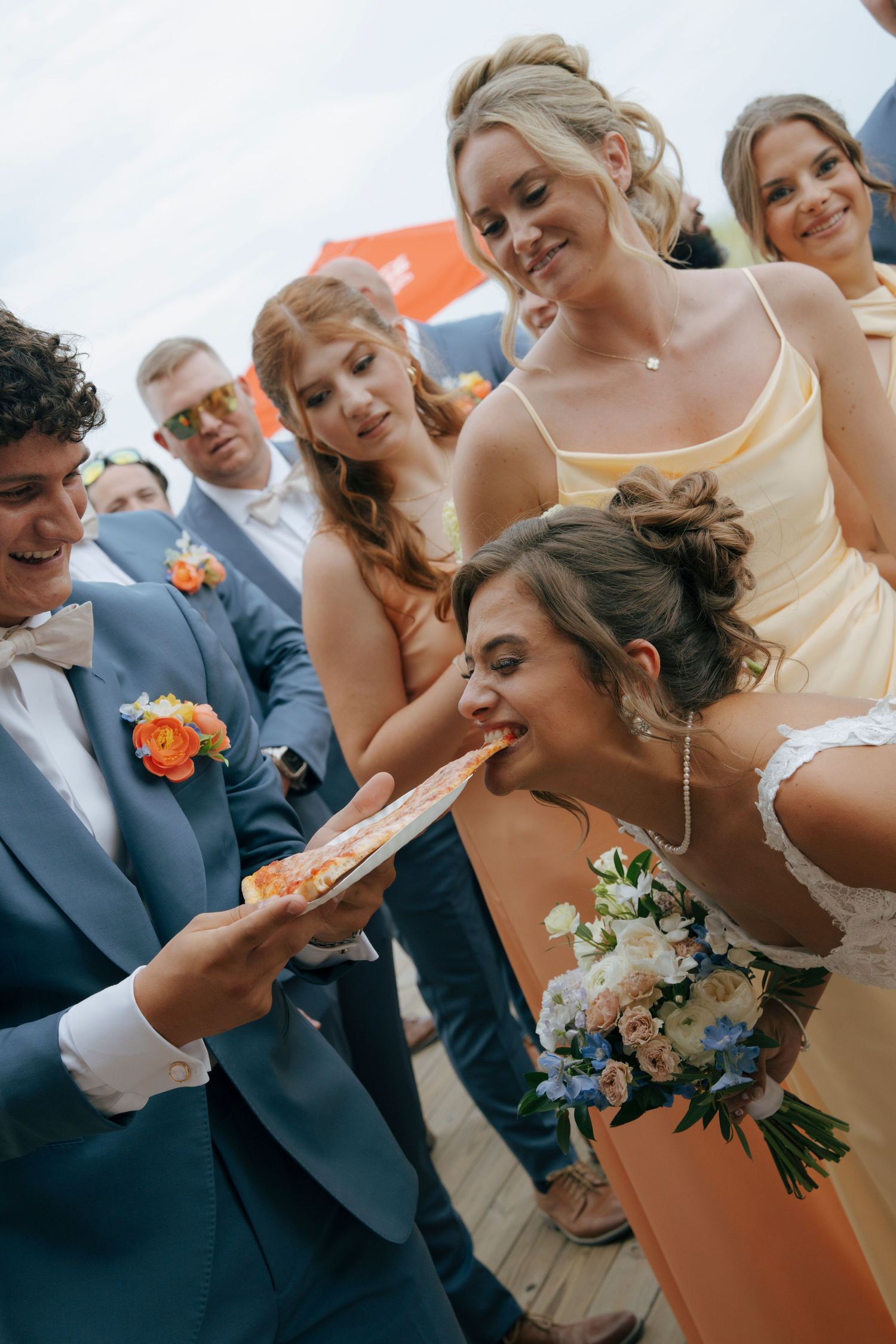 Bride biting food held by groom, surrounded by bridesmaids in yellow dresses at an outdoor wedding.
