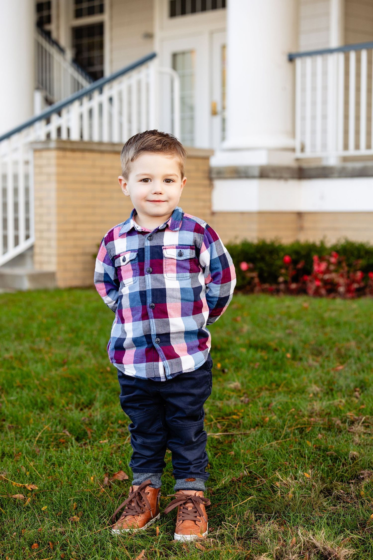 Young boy in plaid shirt and navy pants stands smiling on green lawn in front of white house.