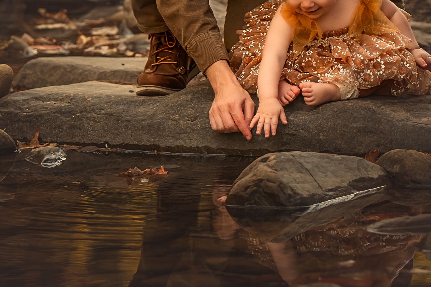 Two children crouch by a rocky stream, gently touching the water with their fingers on an autumn day.