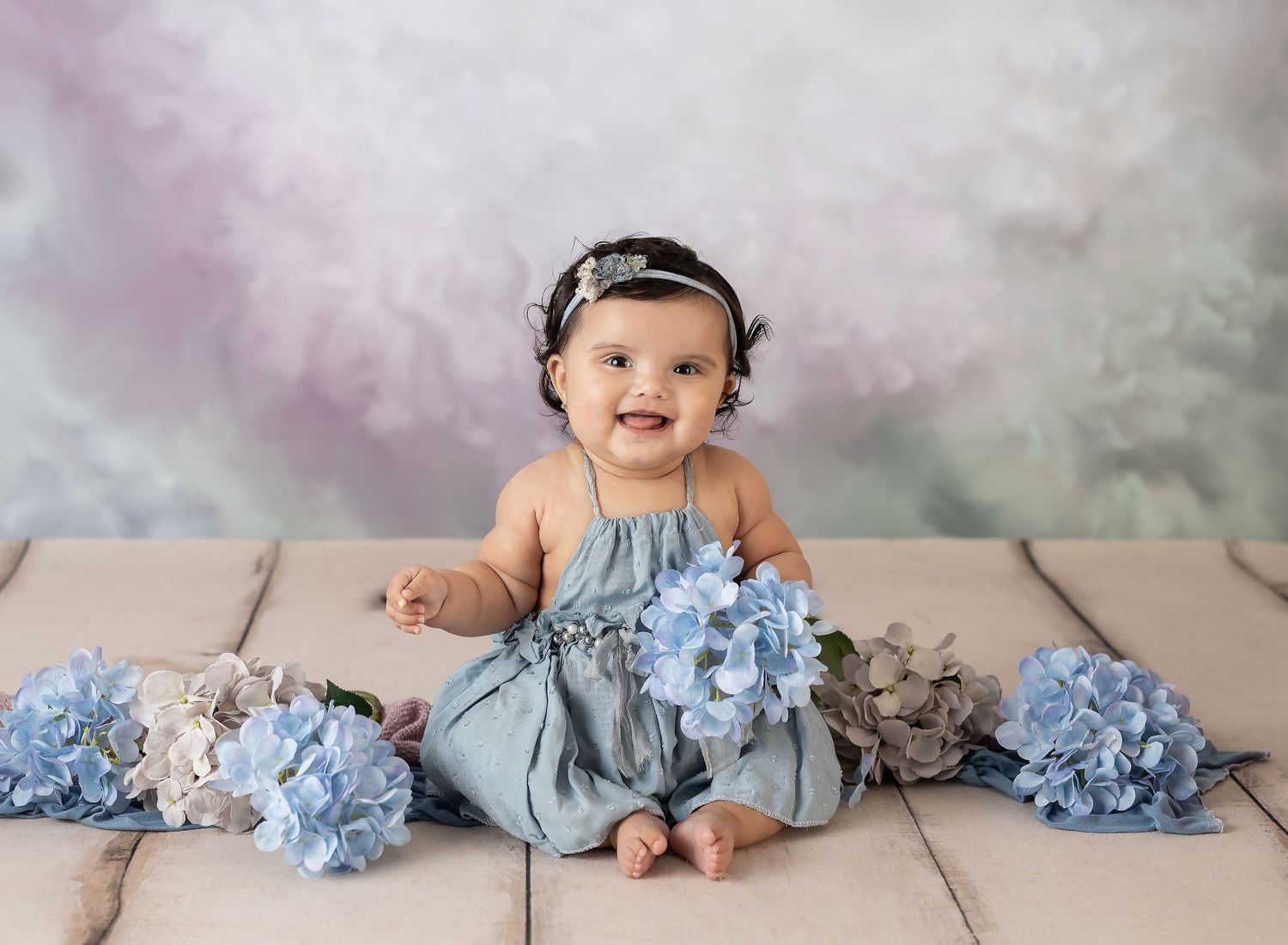 Smiling baby girl in blue romper sitting with hydrangea flowers against a soft pastel backdrop.