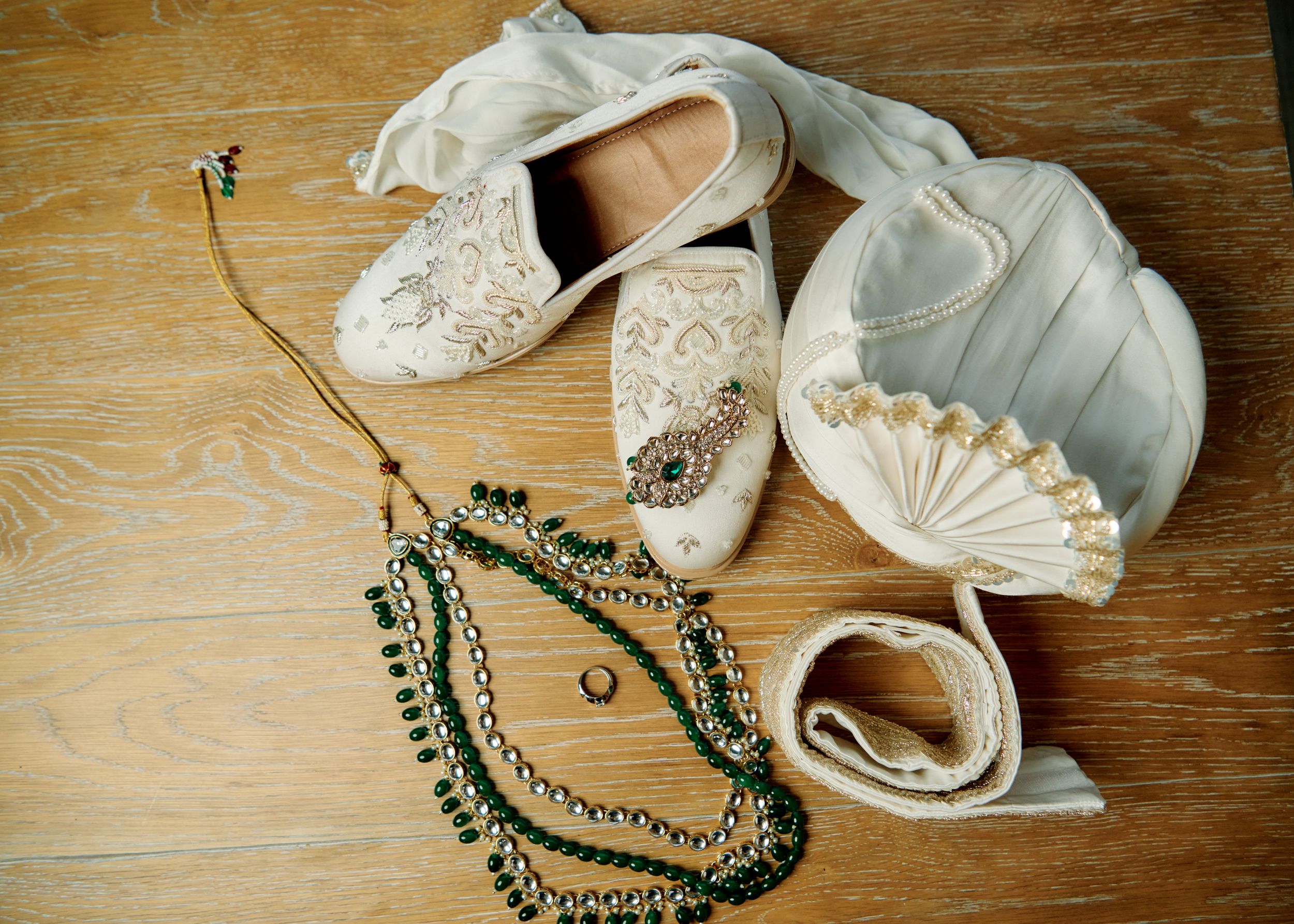 Elegant bridal accessories including lace flats, green beaded necklaces, a gold ring, and a white satin clutch on wood floor.