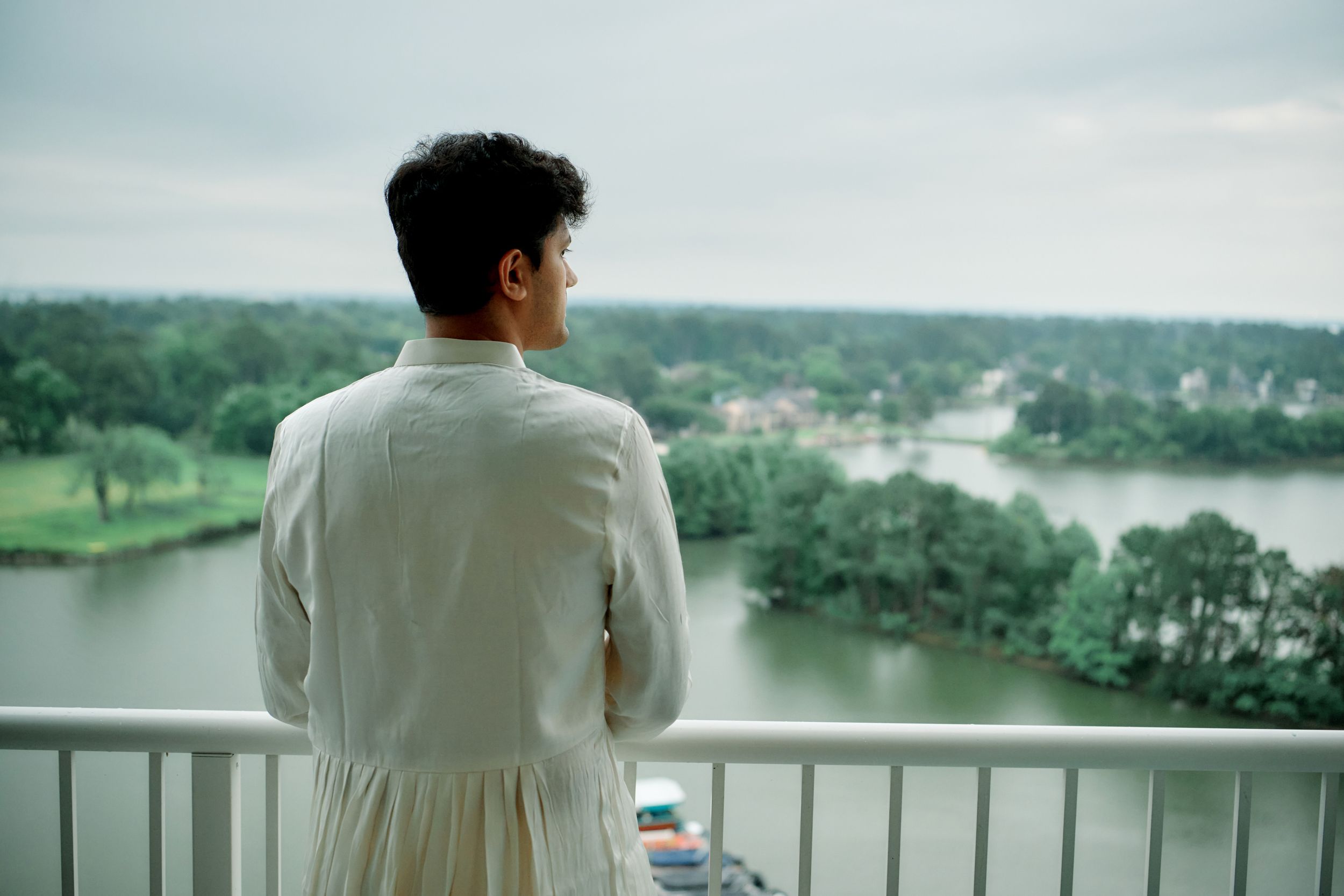 Young man in white outfit gazing at scenic river landscape from high-rise balcony on overcast day.
