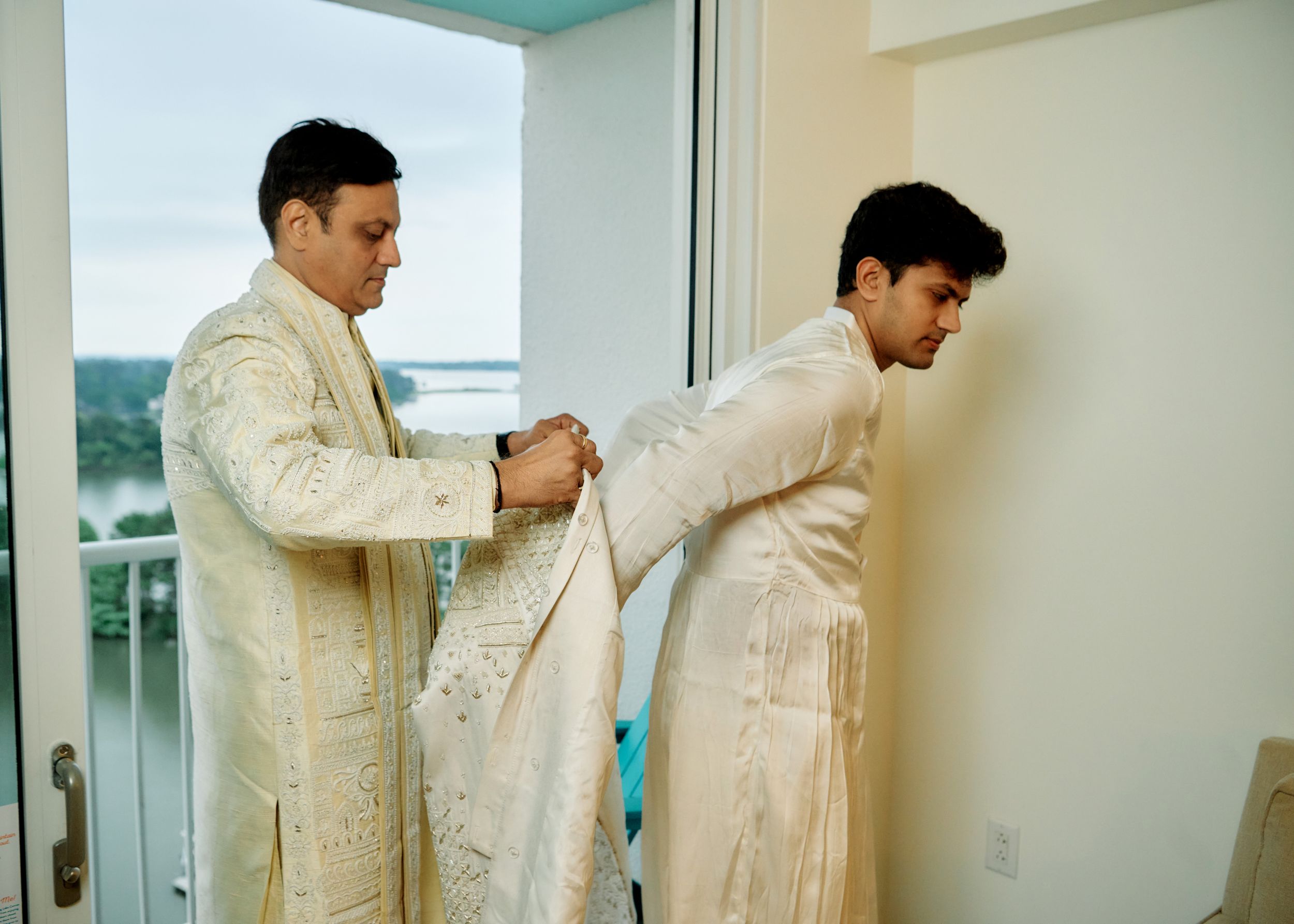 Two men in traditional white embroidered sherwanis assist each other getting dressed near a bright window.