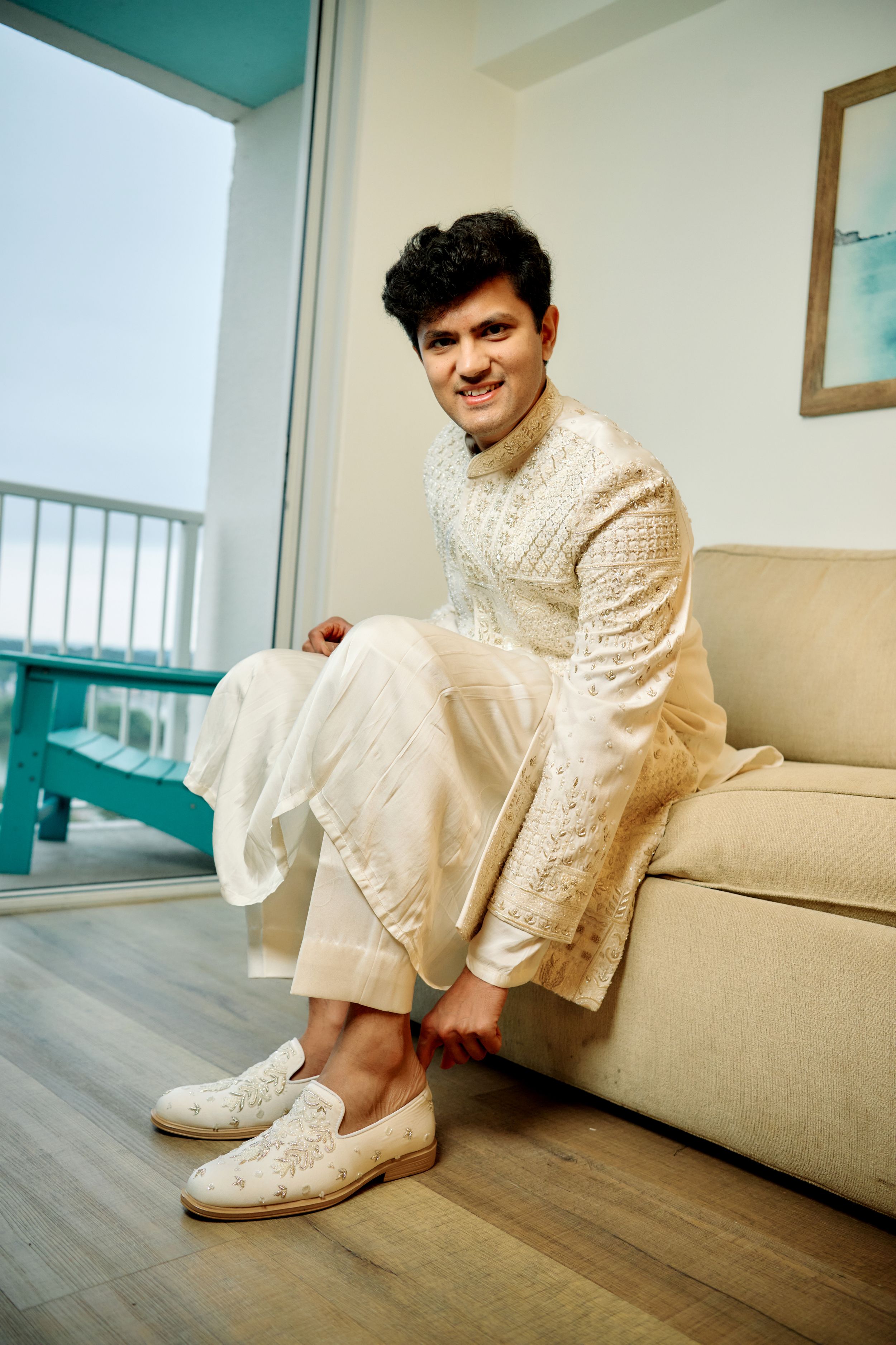 Groom in white embroidered sherwani sitting on beige sofa smiling, wearing embroidered mojari shoes.